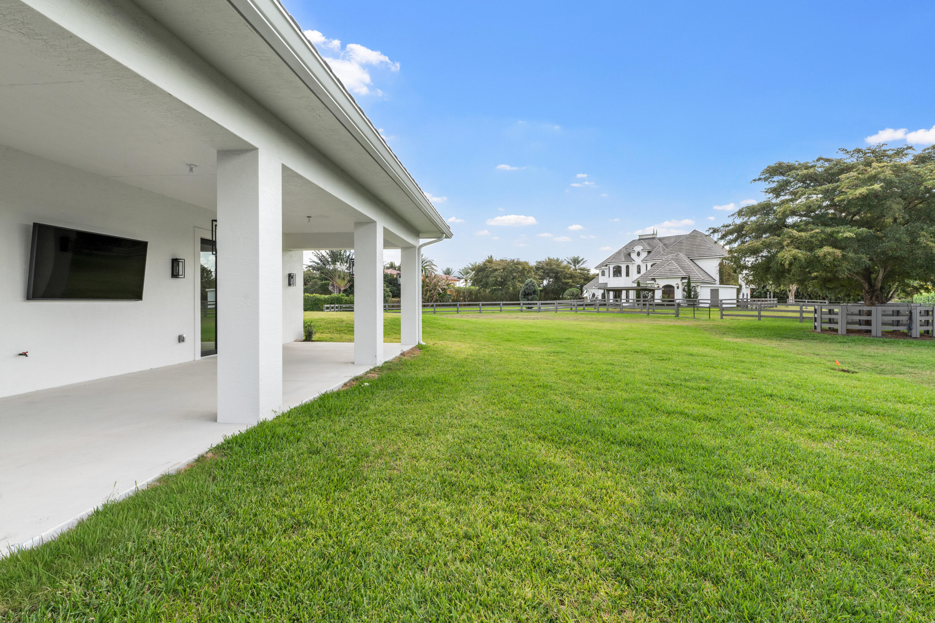 Covered patio beside a broad green lawn, with a fenced white house visible across the yard.