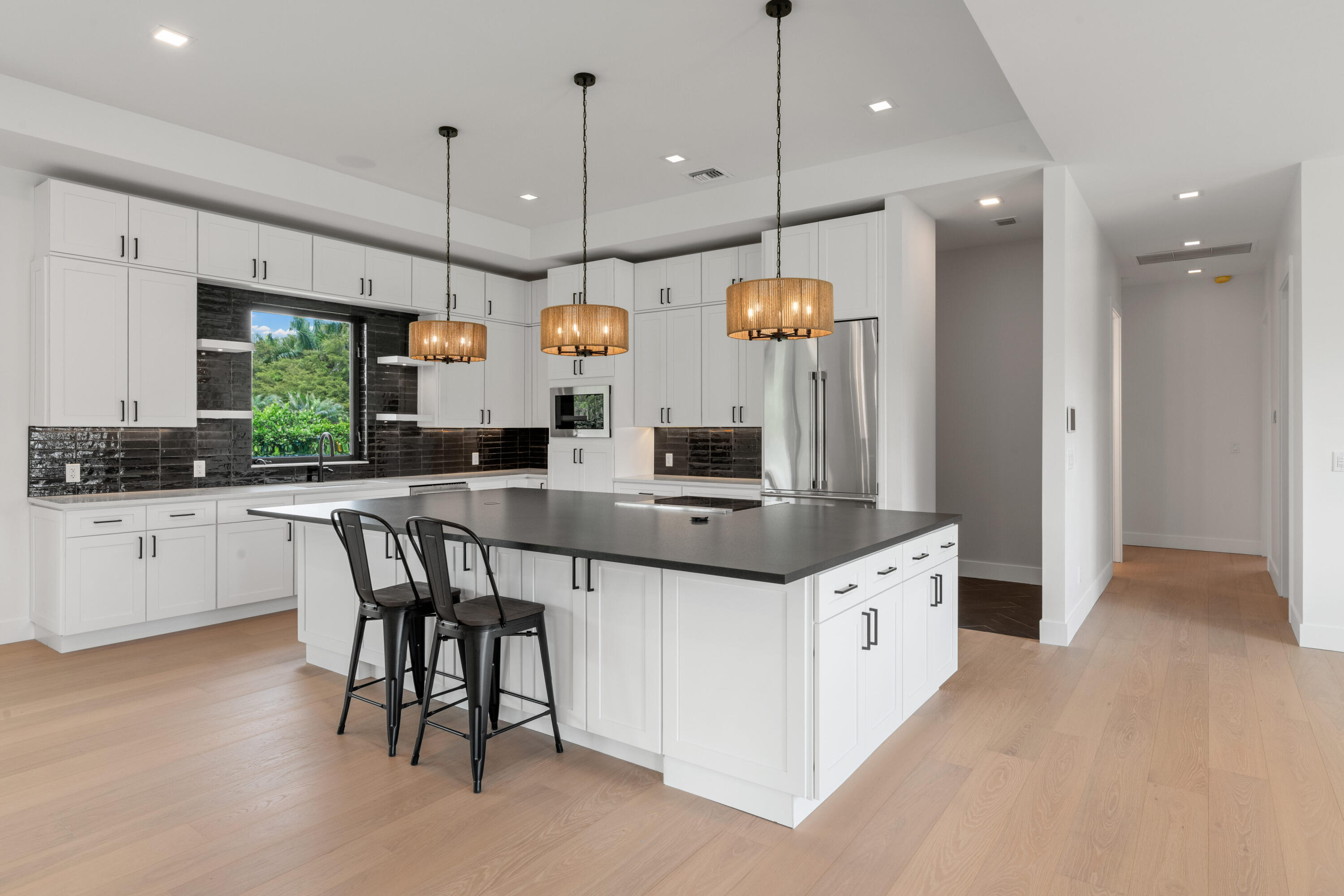 Modern white kitchen with a large dark island, pendant lights, and a window overlooking greenery.