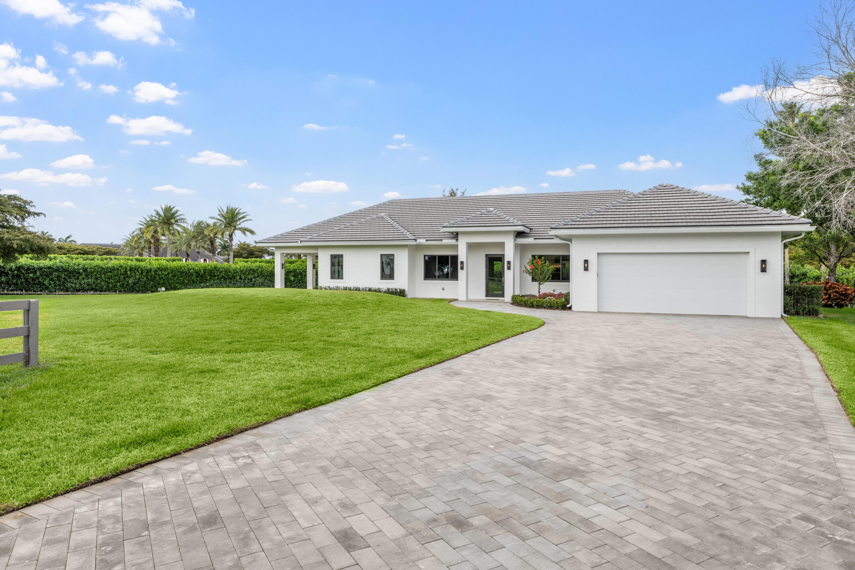 Single-story white home with a gray tiled roof, attached garage, broad paver driveway, and spacious lawn.