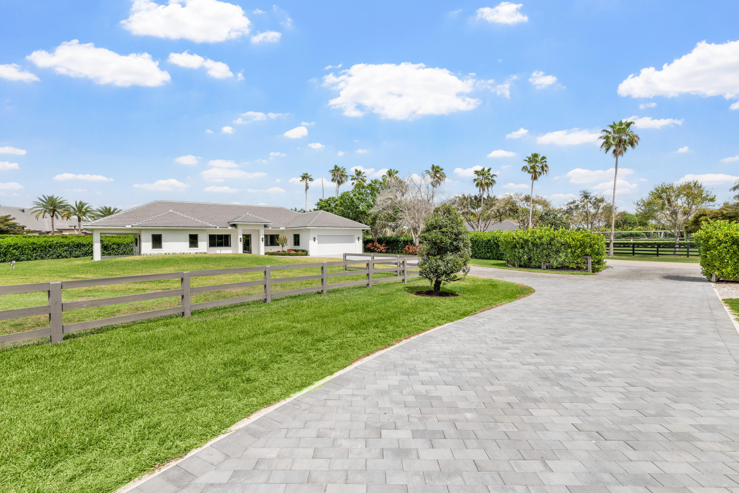 Single-story white house with a broad paver driveway, lawn, and palm trees.