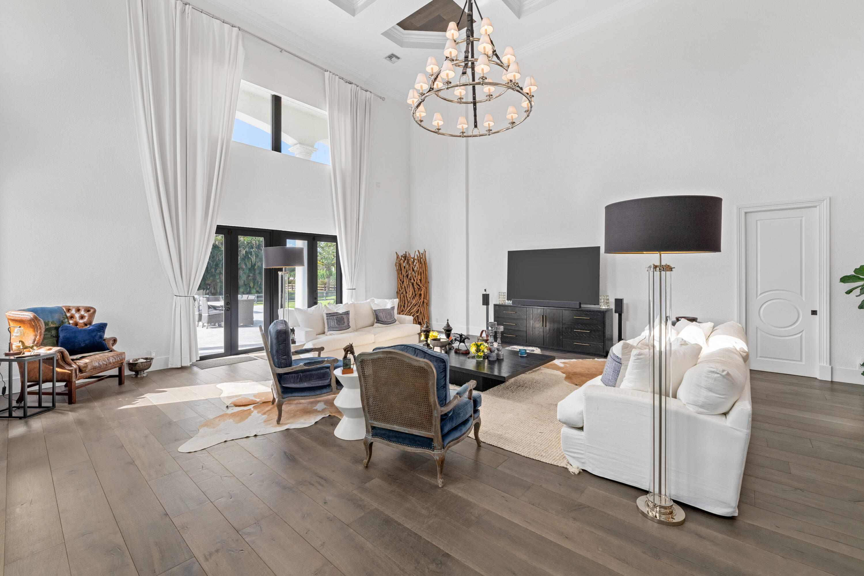 Living room with soaring ceilings, a chandelier, tall curtained windows, white sofas, and blue accent chairs.