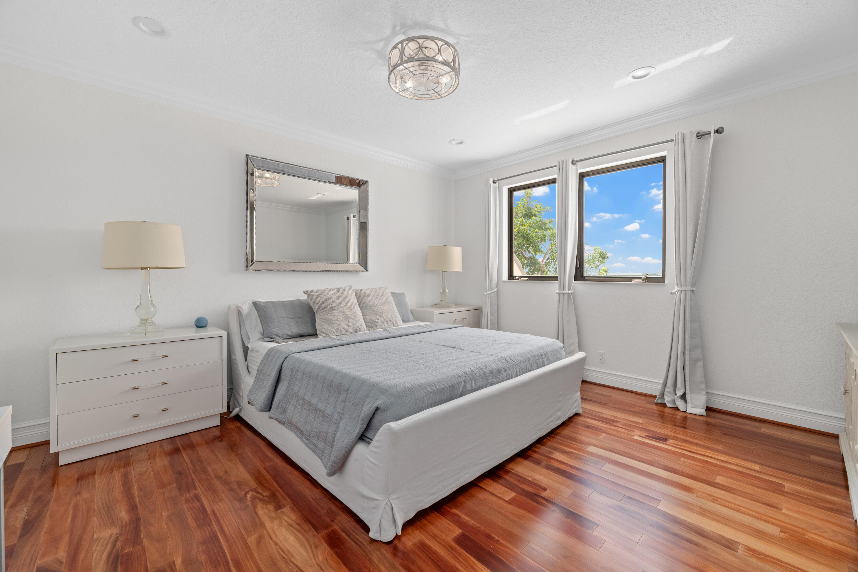 Furnished bedroom with a gray bed, warm hardwood floors, and two windows overlooking trees.