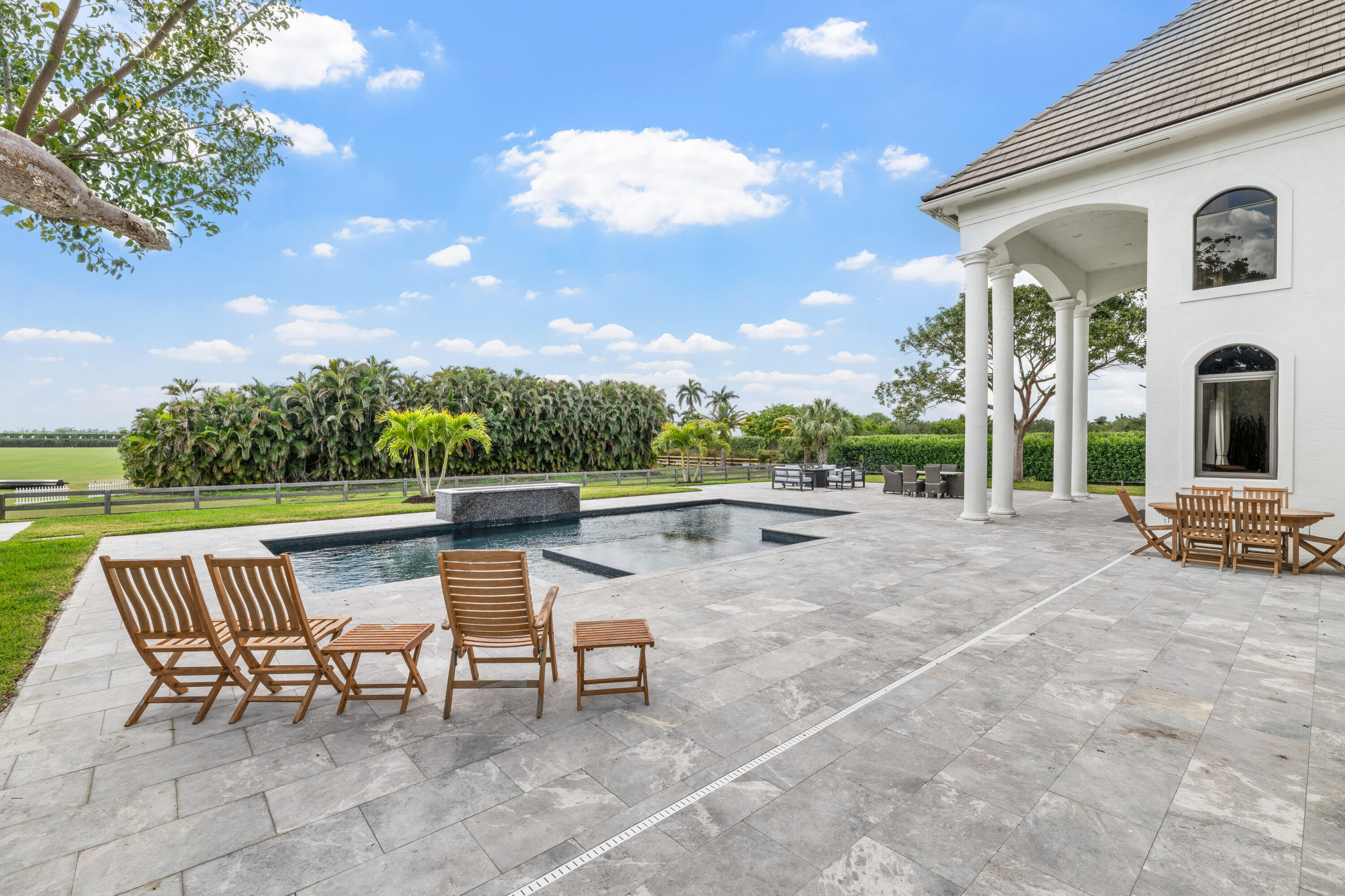 Outdoor pool and stone terrace with wooden lounge chairs beside a covered, columned porch.