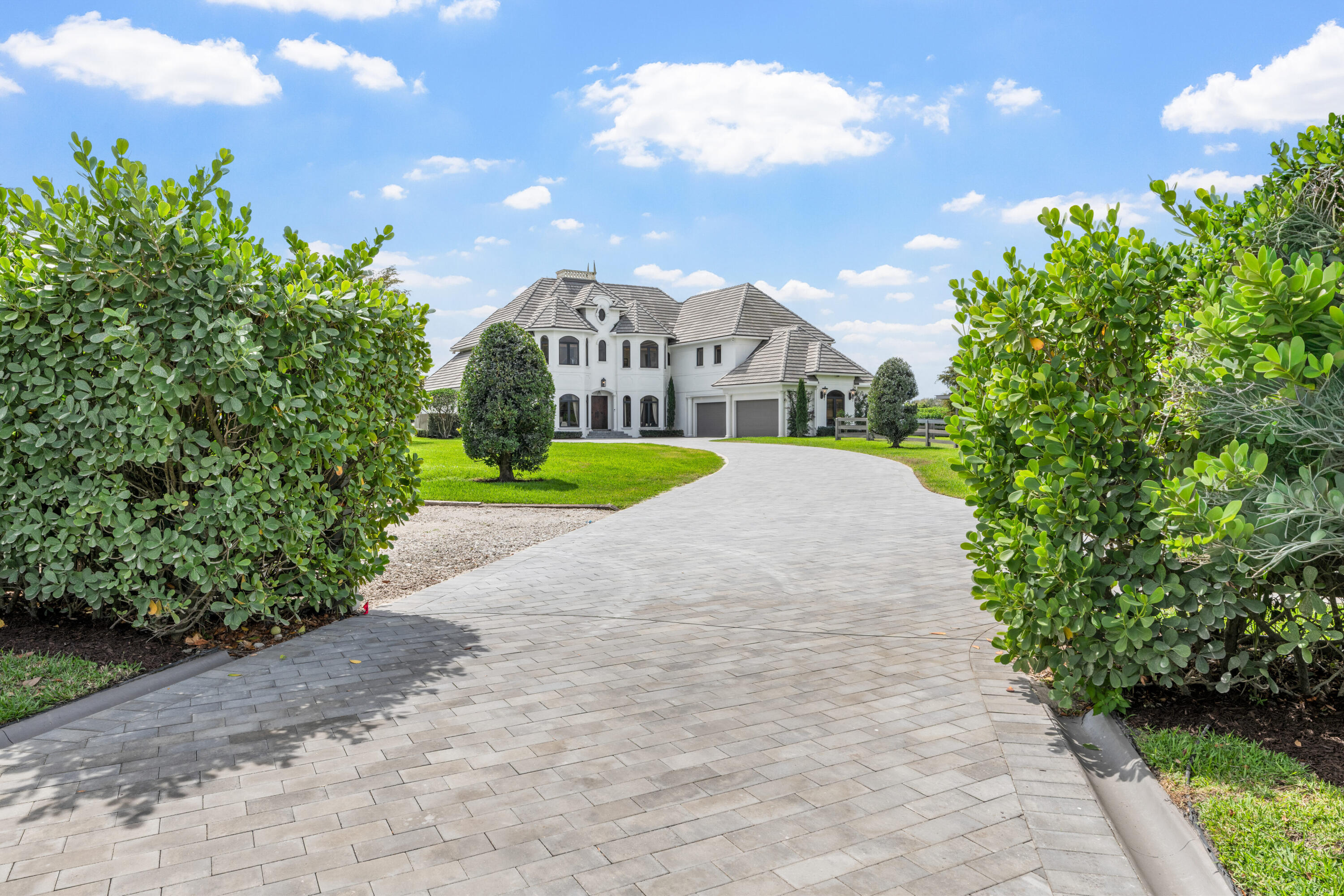 Two-story white home with a gray tiled roof, framed by hedges beyond a curved paver driveway.
