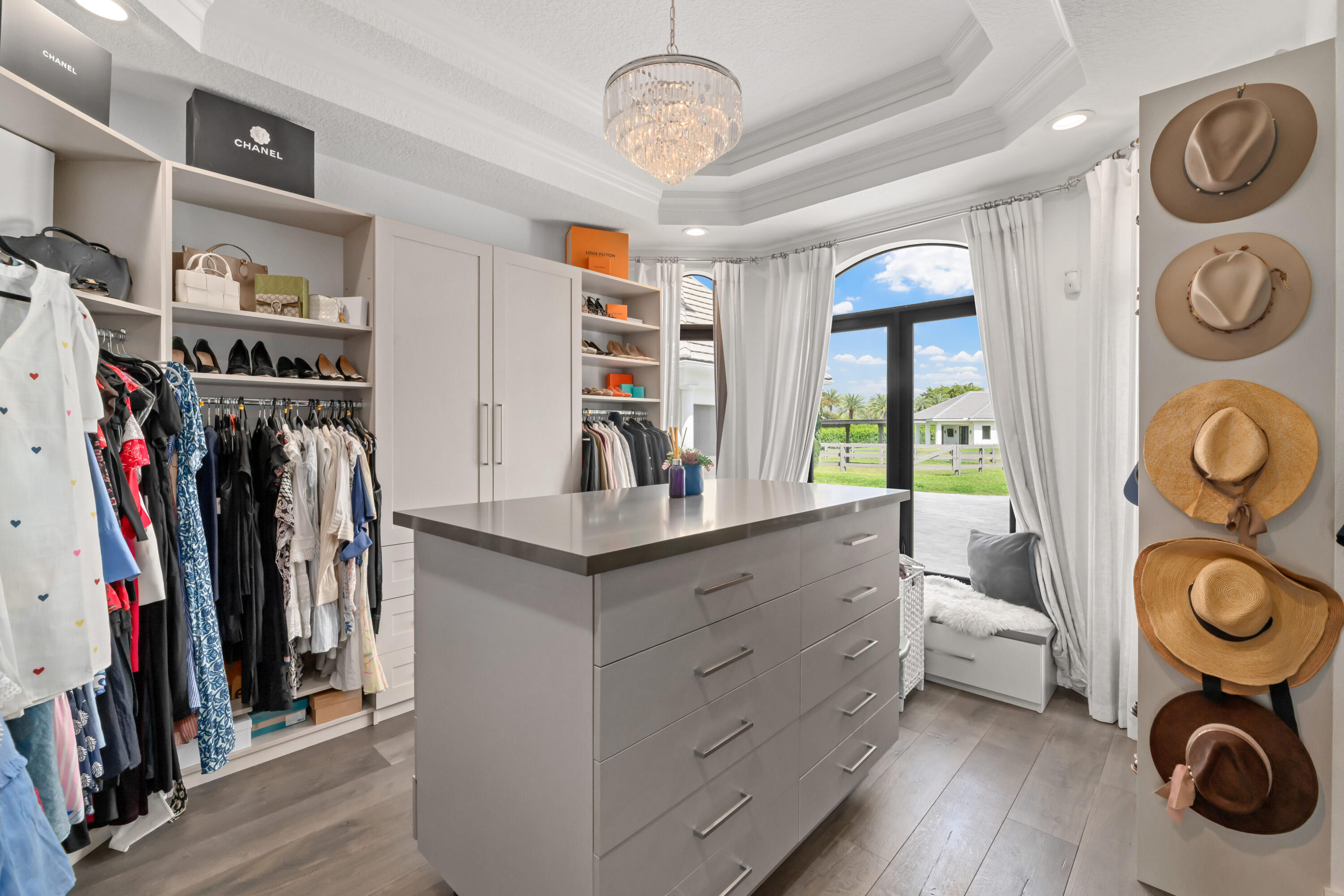 Walk-in closet with a central drawer island, built-in clothing shelves, and hats displayed beside an arched window.