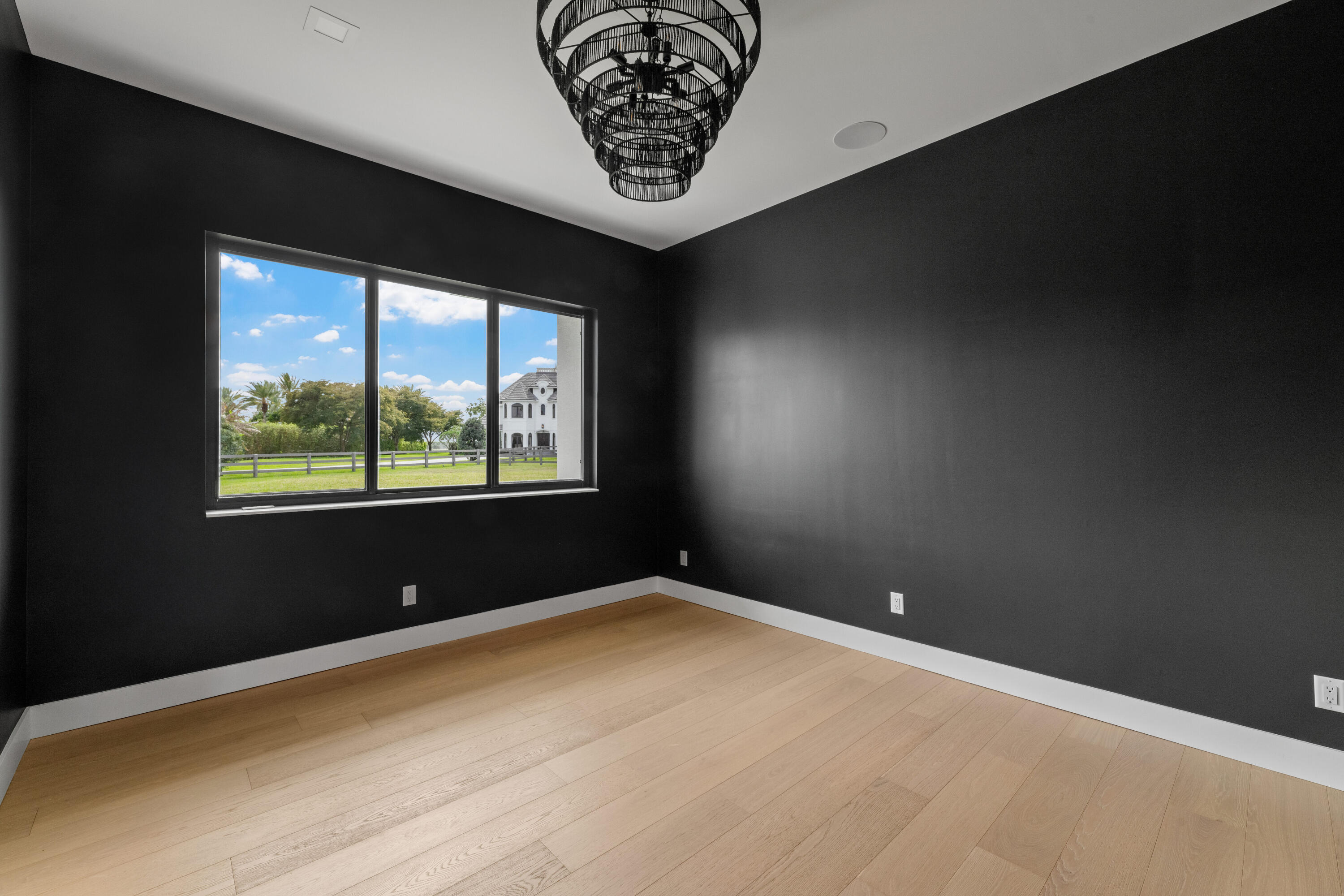 Unfurnished room with black walls, pale oak floors, a large chandelier, and a wide window overlooking a lawn.