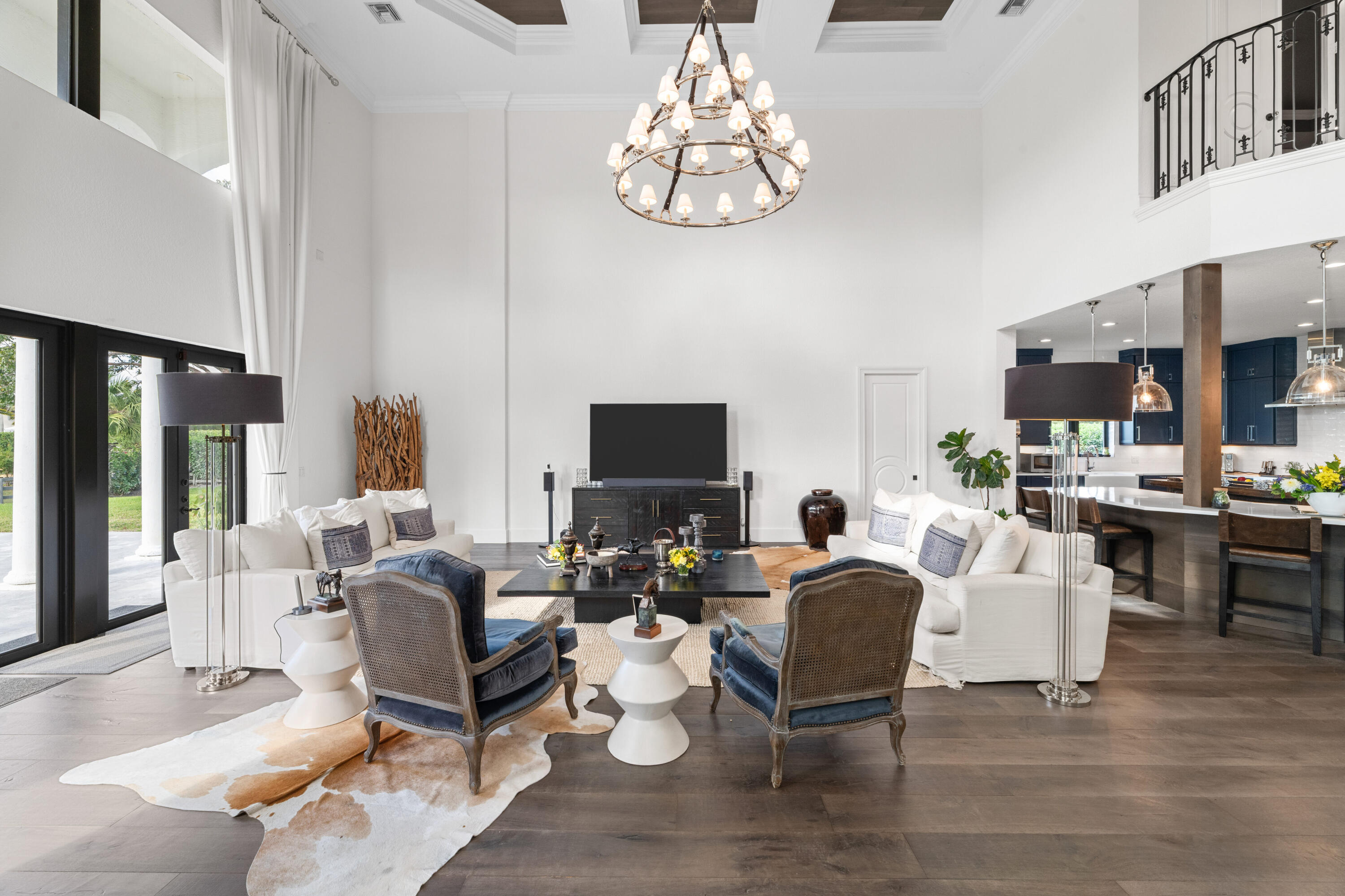 Double-height living room with white sofas, blue chairs, a large chandelier, and an open kitchen.