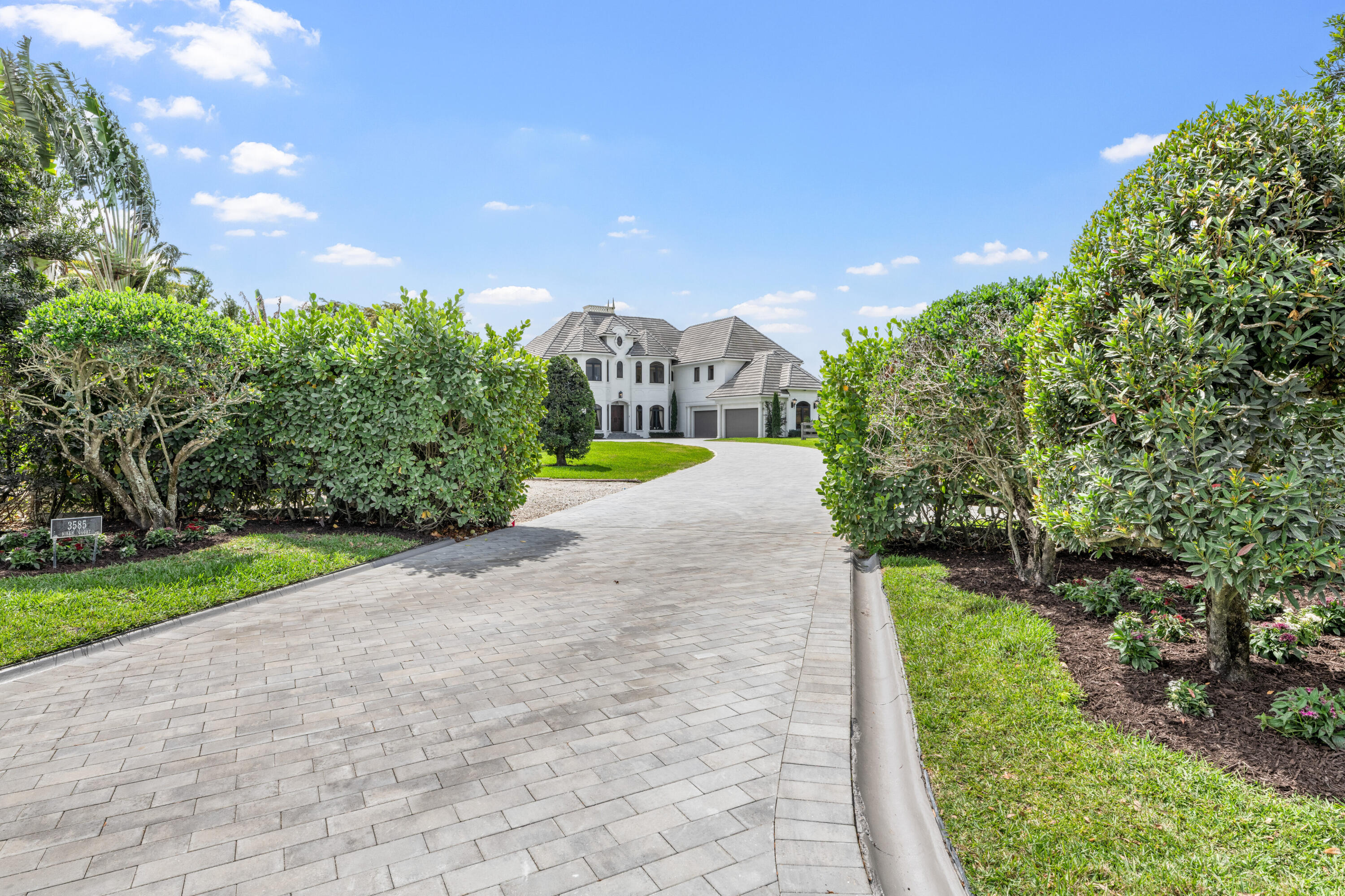 White two-story home with a tiled roof, framed by hedges at the end of a wide paver driveway.