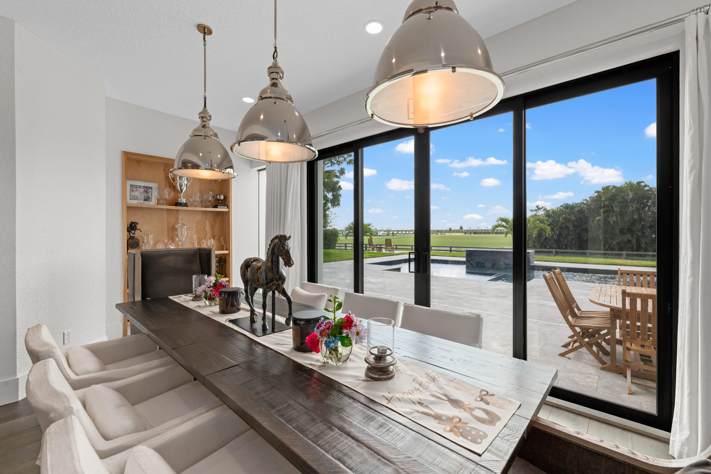 Dining room with long wood table, pendant lights, and pool view through glass doors.