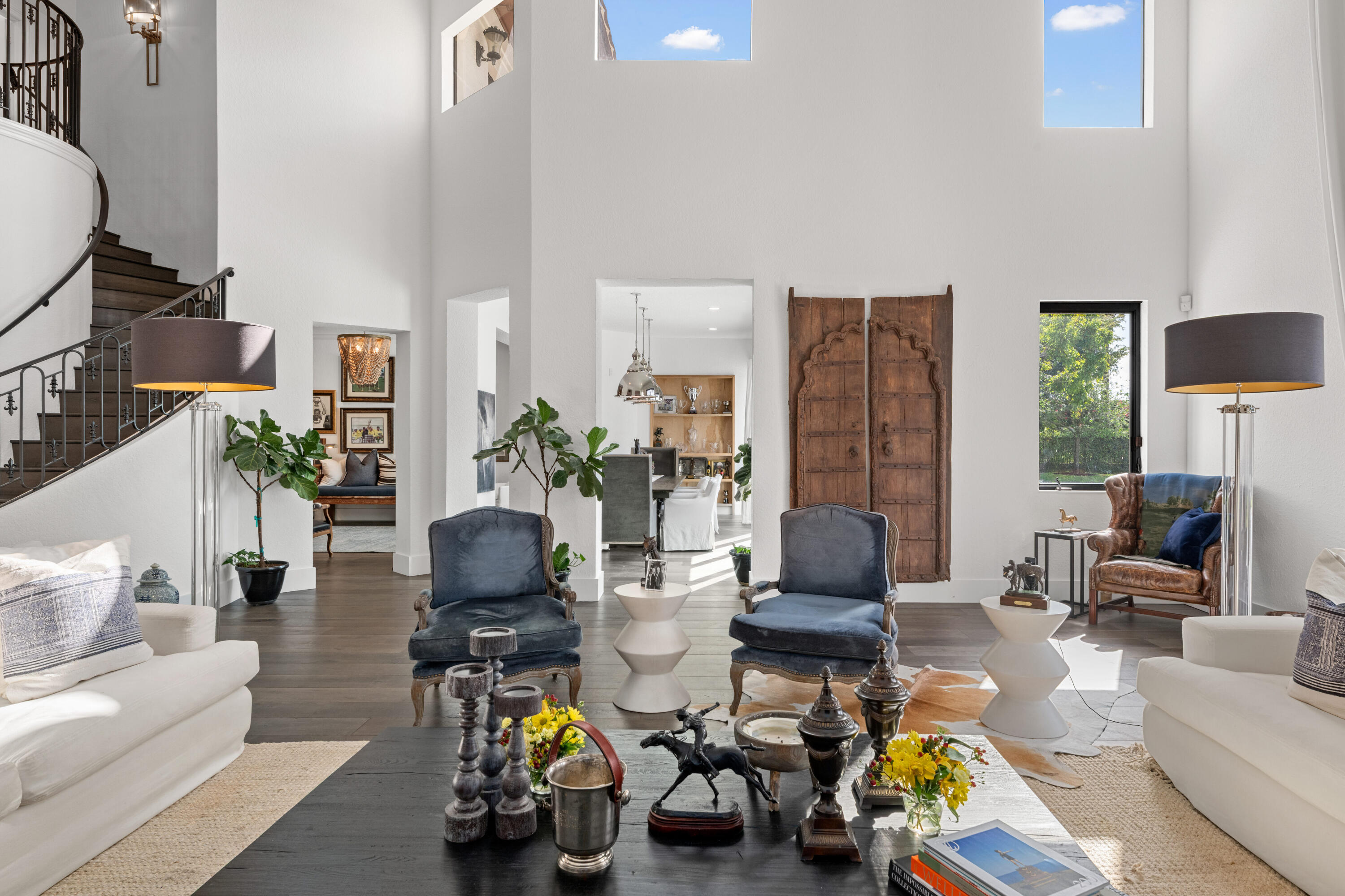 Double-height living room with a curved staircase, blue armchairs, and rustic carved wooden doors.