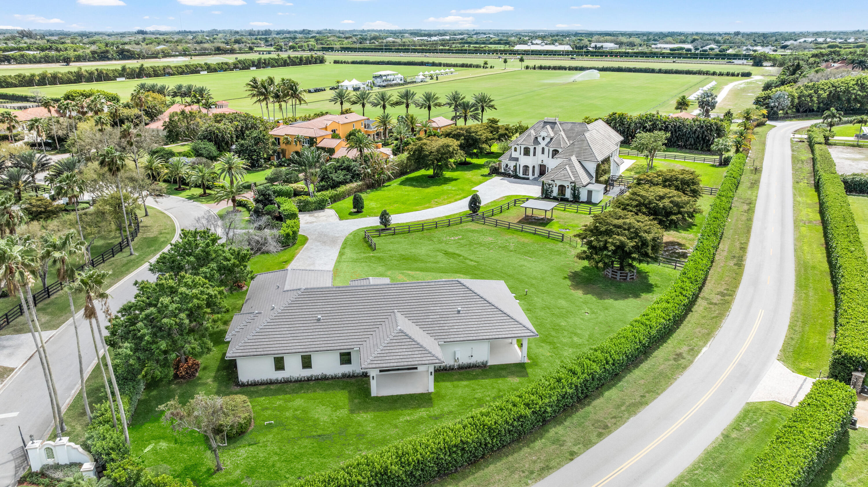 Aerial view of a tile-roofed home beside expansive polo fields, with palm-lined grounds and a neighboring estate.