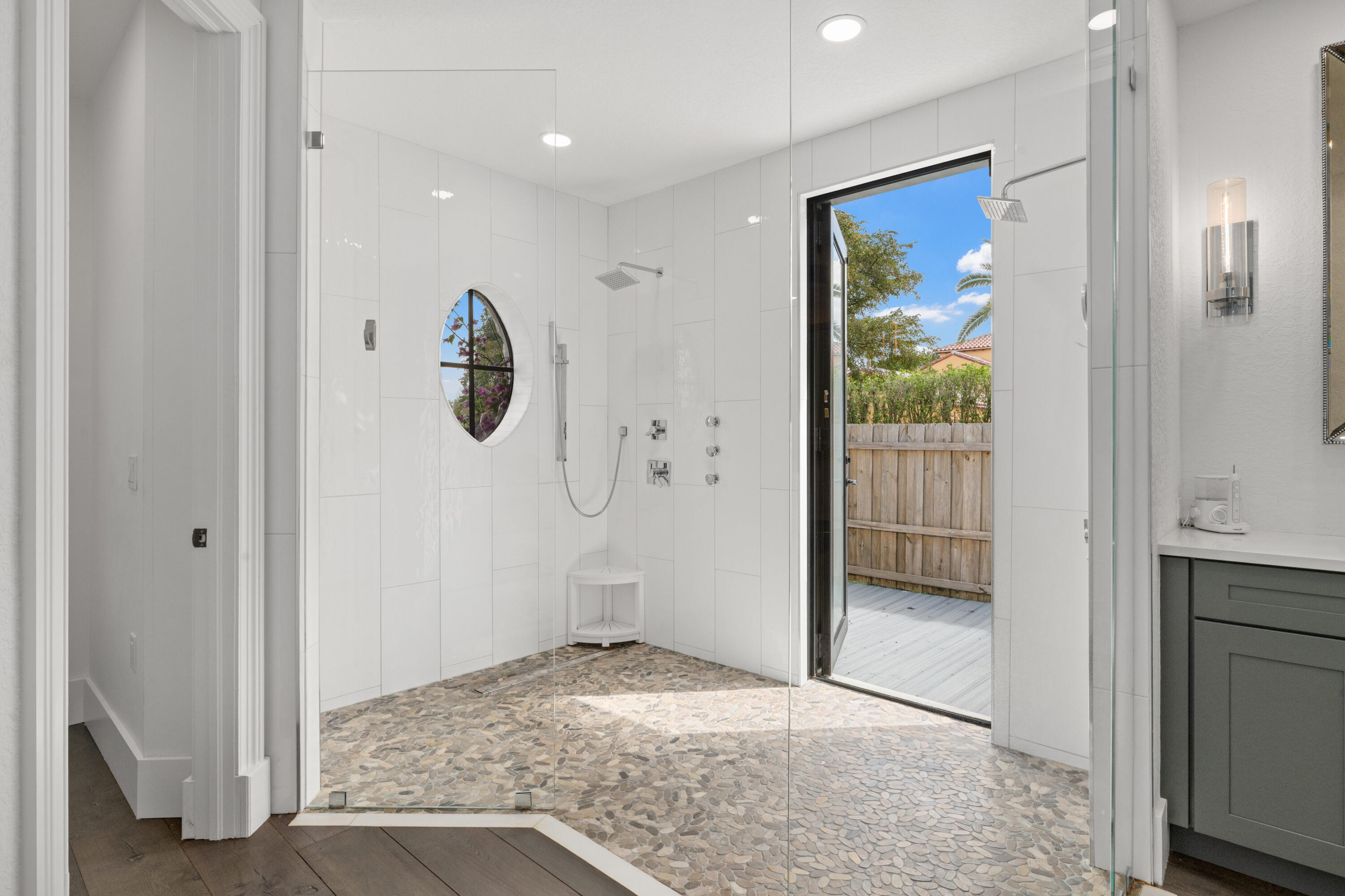 Modern walk-in shower with pebble flooring, oval window, and an open door to a private deck.
