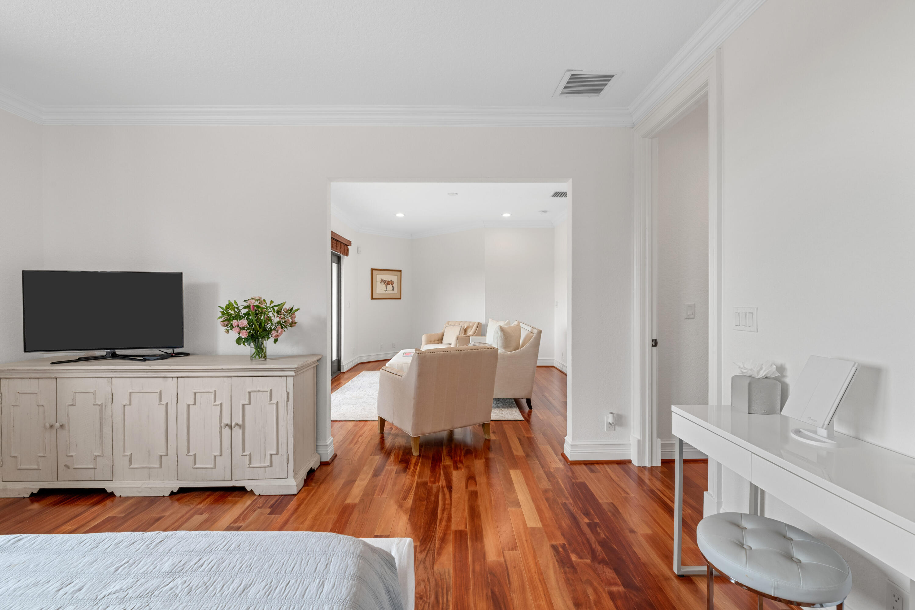 Bedroom with a white bed and TV cabinet, opening onto a sitting area with cream armchairs.