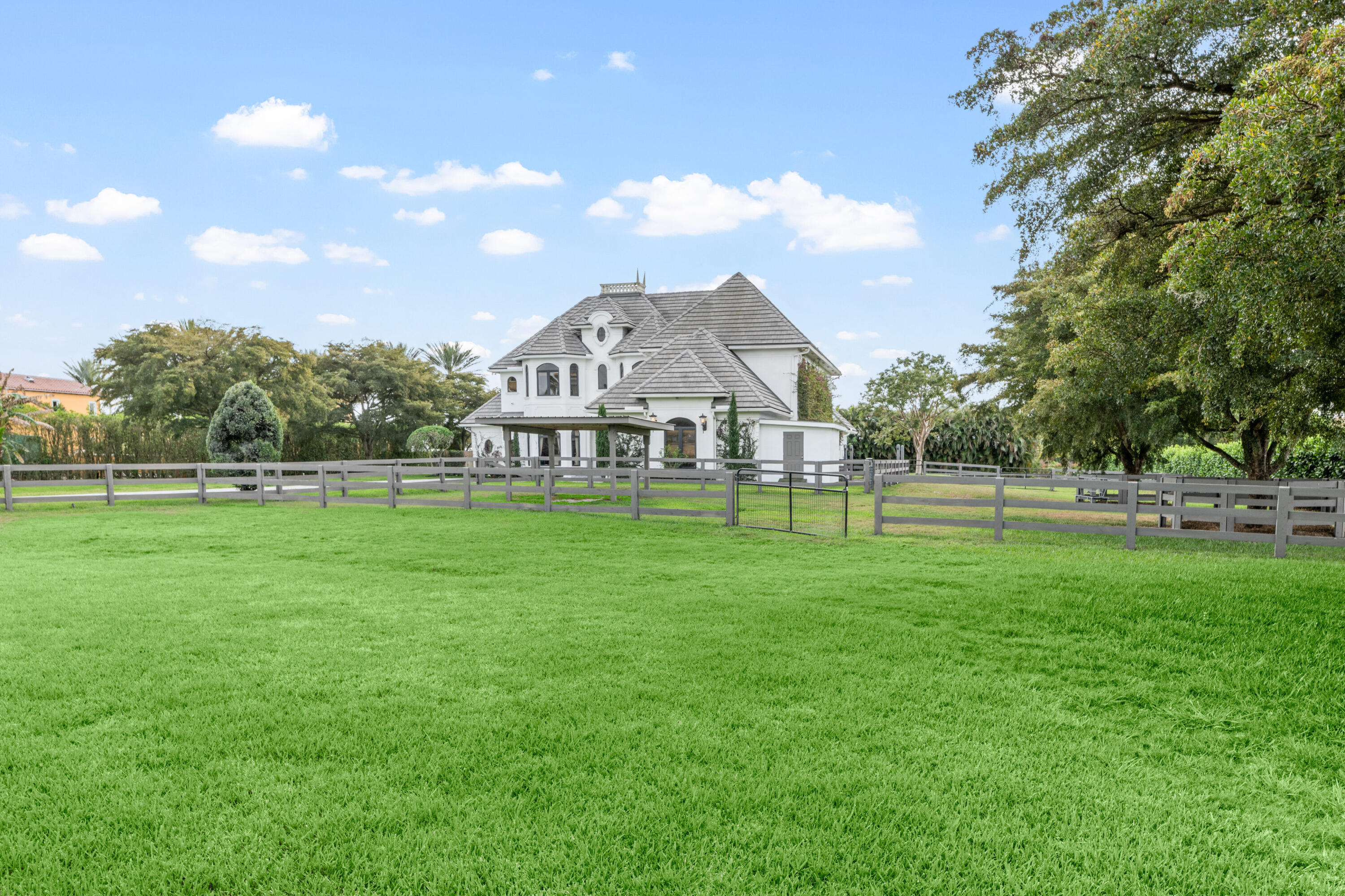 White multi-story home beyond a broad fenced lawn, framed by mature trees.