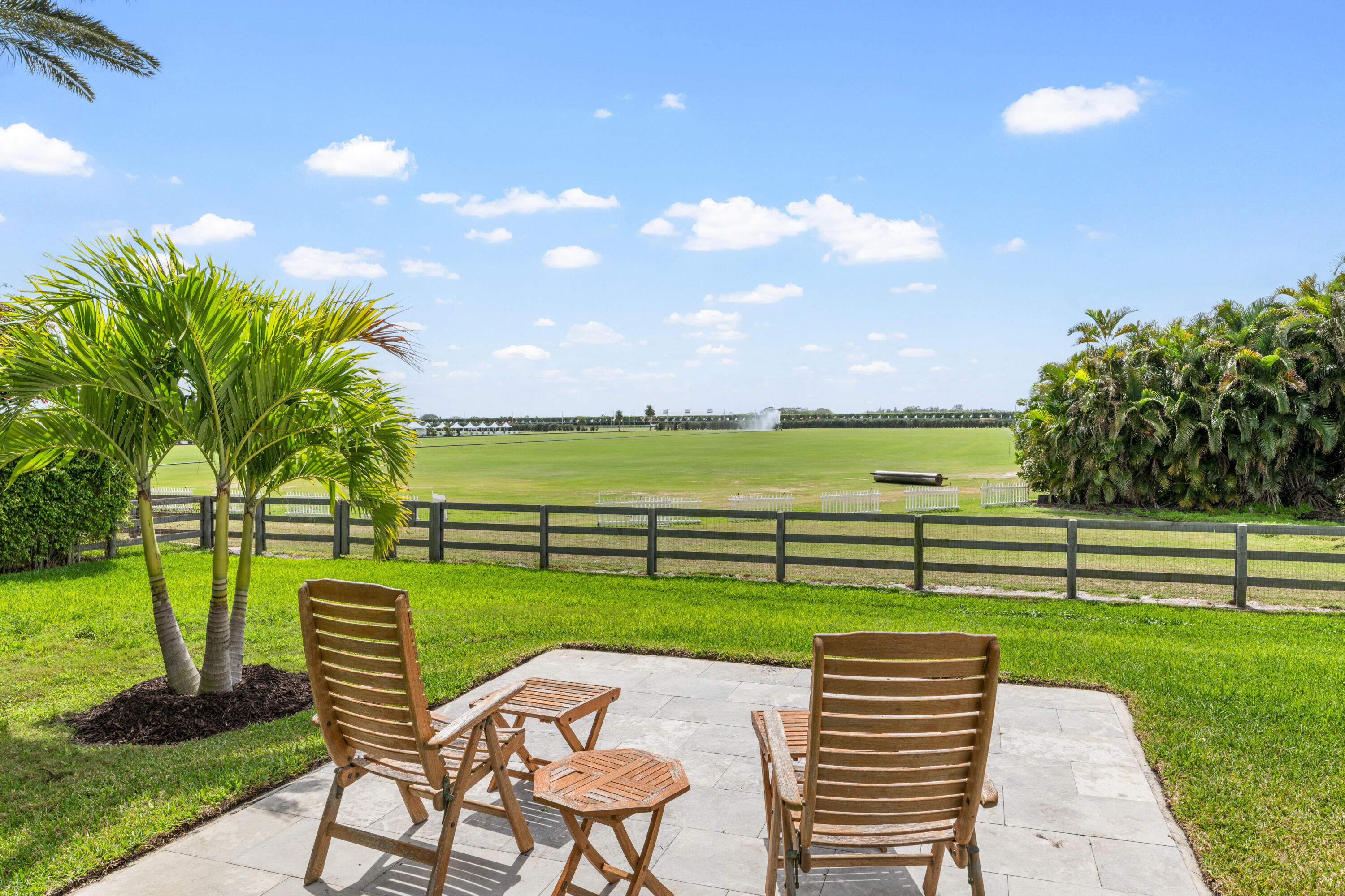 Stone patio with wooden seating overlooking a fenced polo field, framed by palm trees.