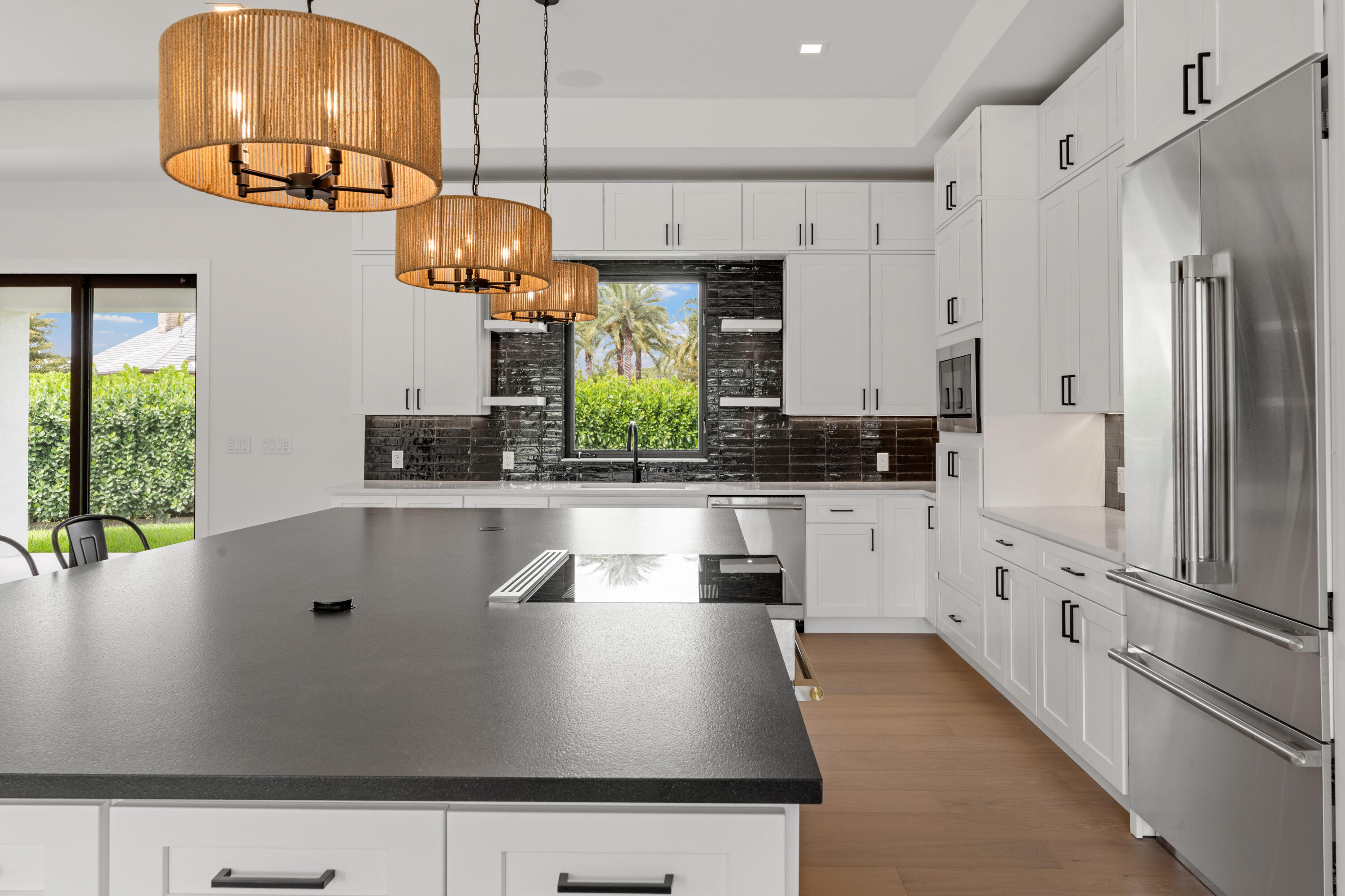 Modern white kitchen with a large dark island, woven pendant lights, and a window overlooking palms.