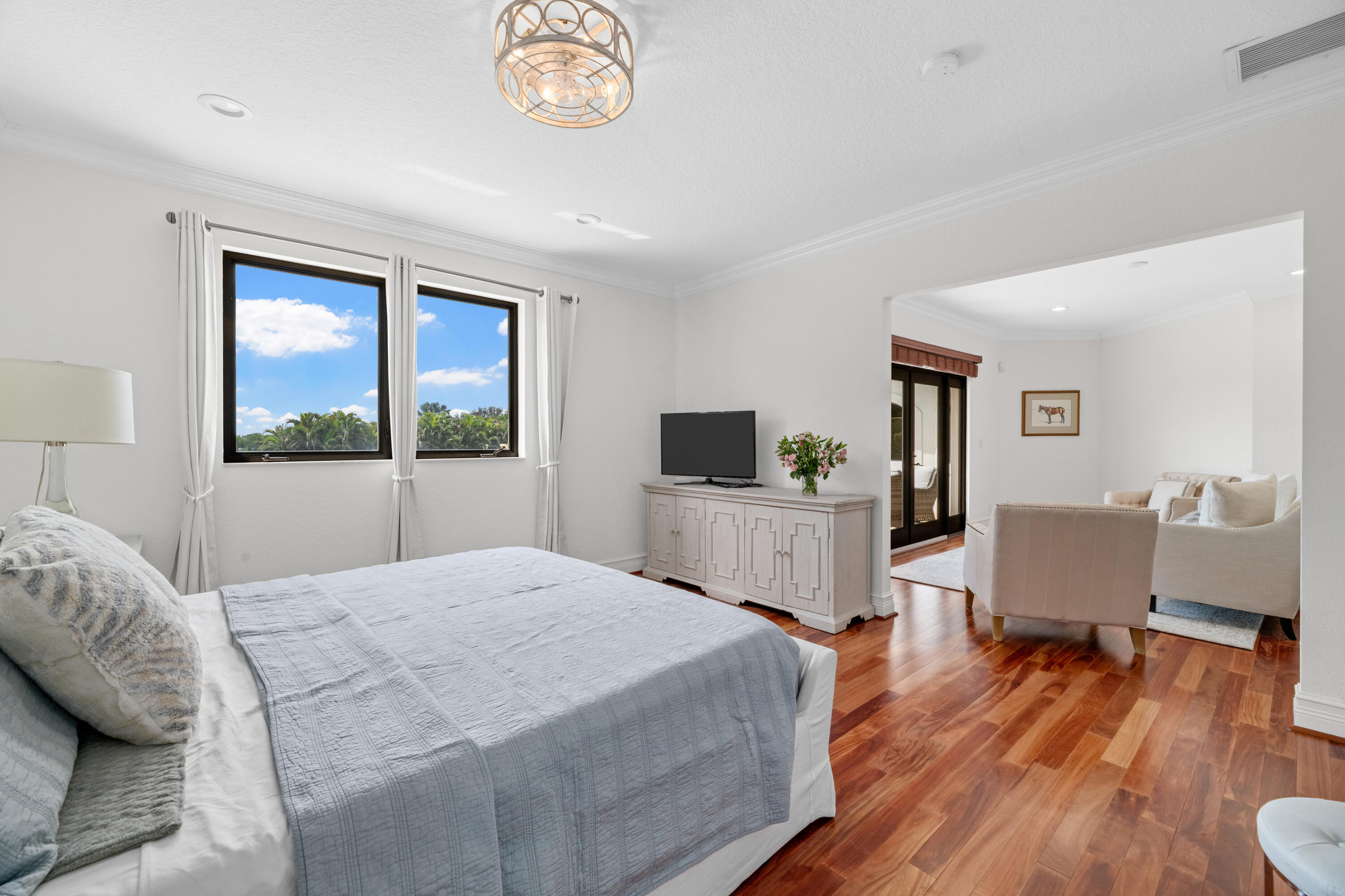 Bright bedroom with a gray quilted bed, wood floors, and a window overlooking palm trees.