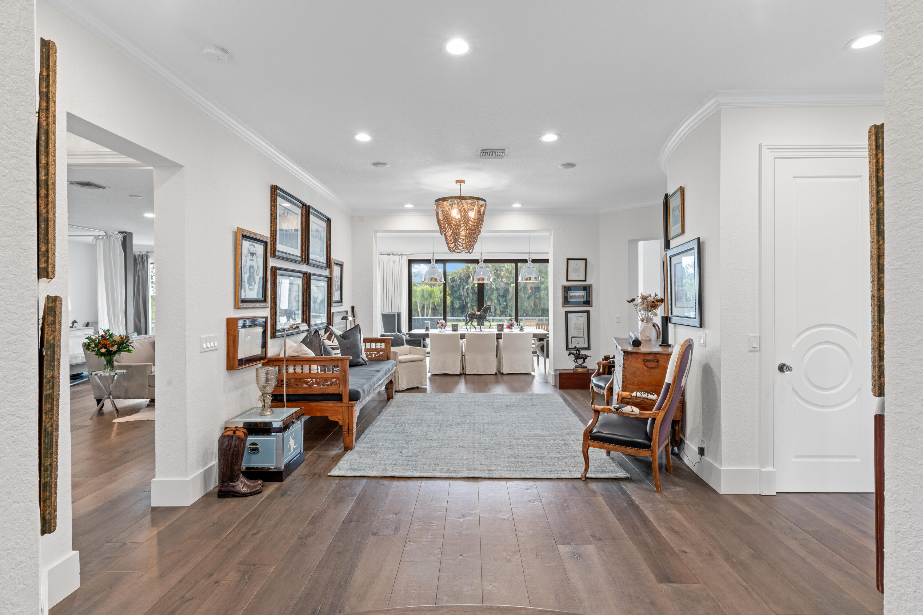Furnished living room opens to a dining area with a chandelier and tropical greenery beyond the windows.