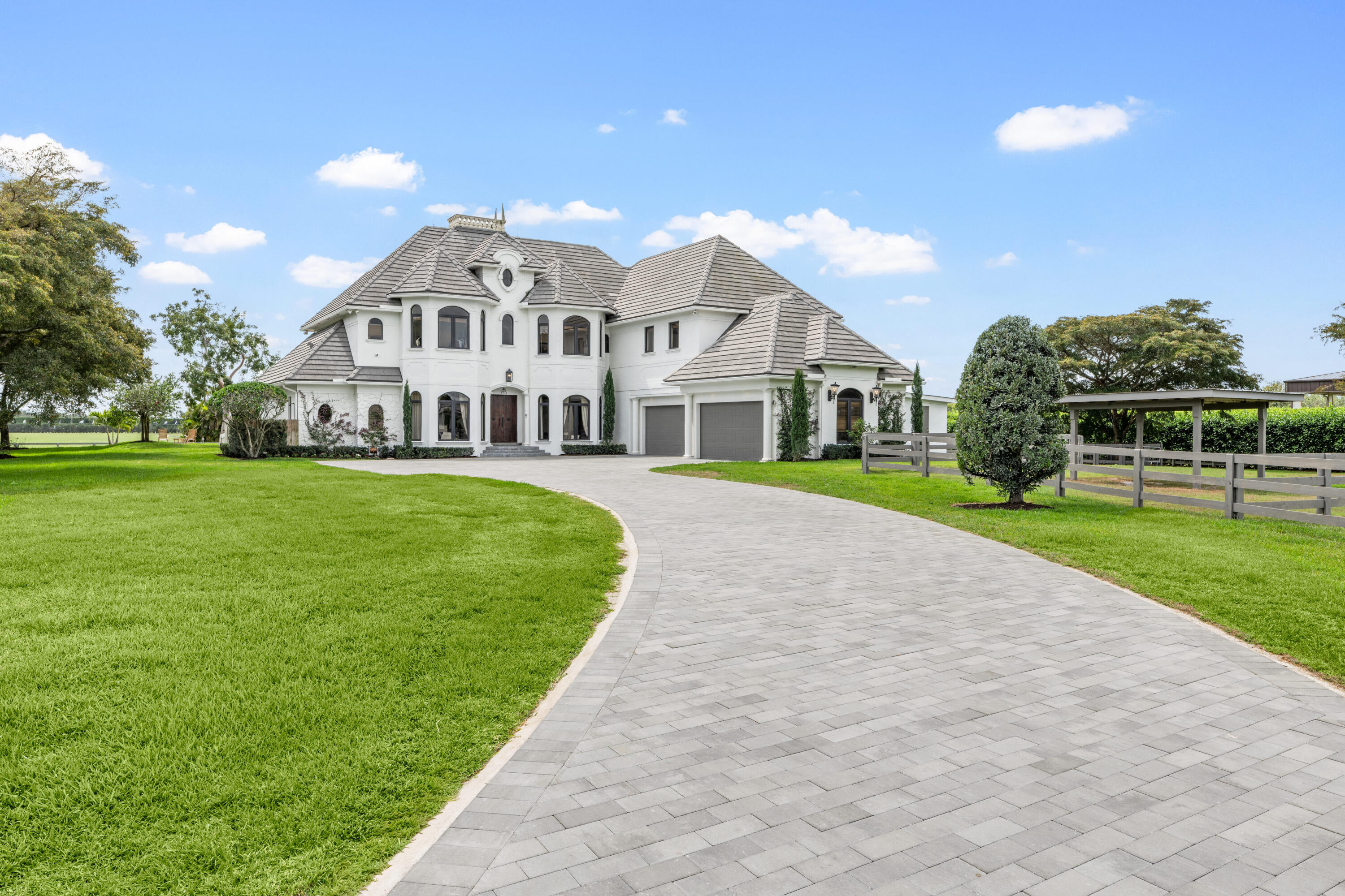 White two-story home with gray tiled roofs, curved paver driveway, expansive lawn, and fenced grounds.