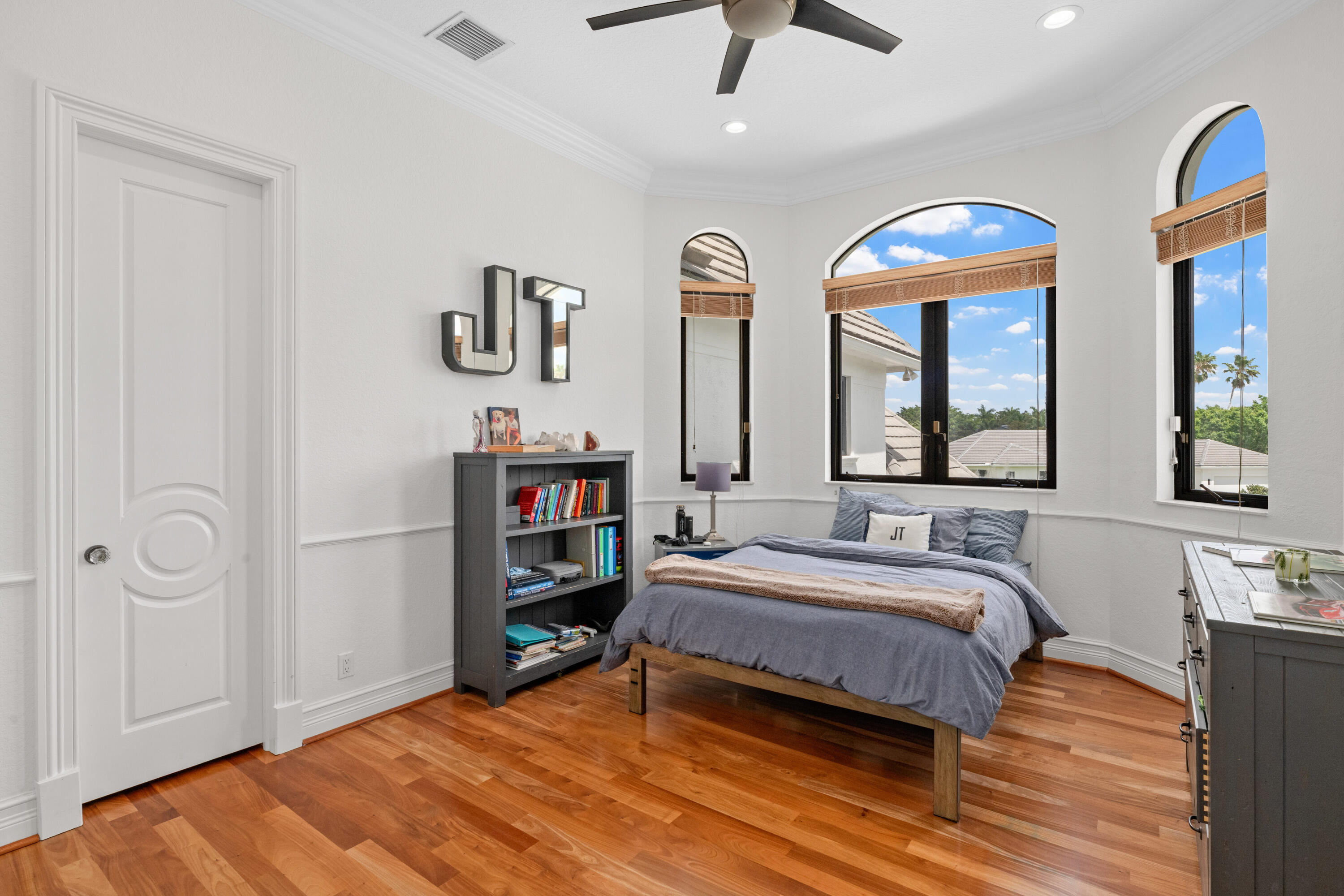 Bedroom with a gray bed, warm wood floors, and three arched windows overlooking palm trees.