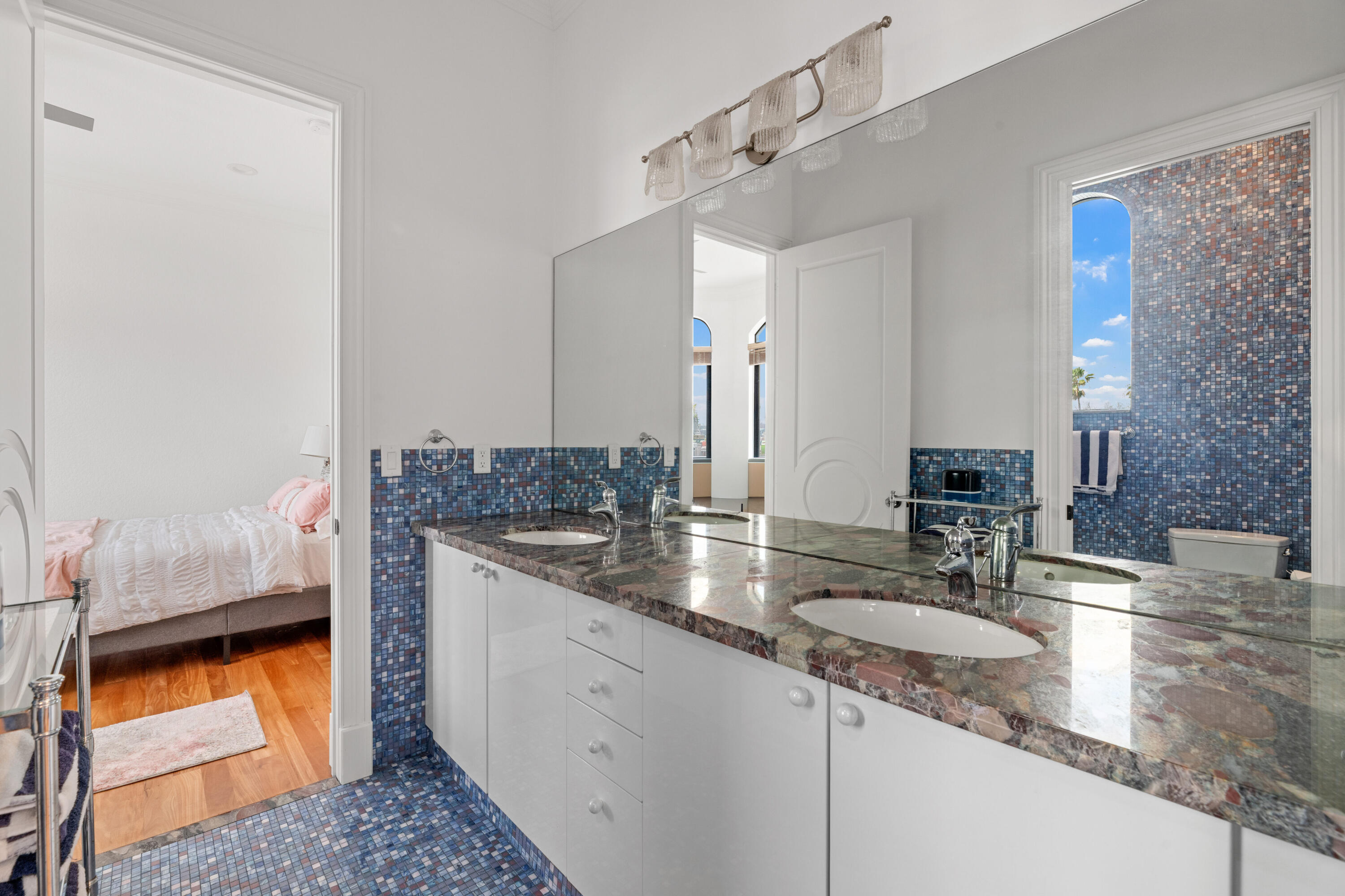 Bathroom with a double-sink stone vanity and blue mosaic tile, opening to a pink-accented bedroom.