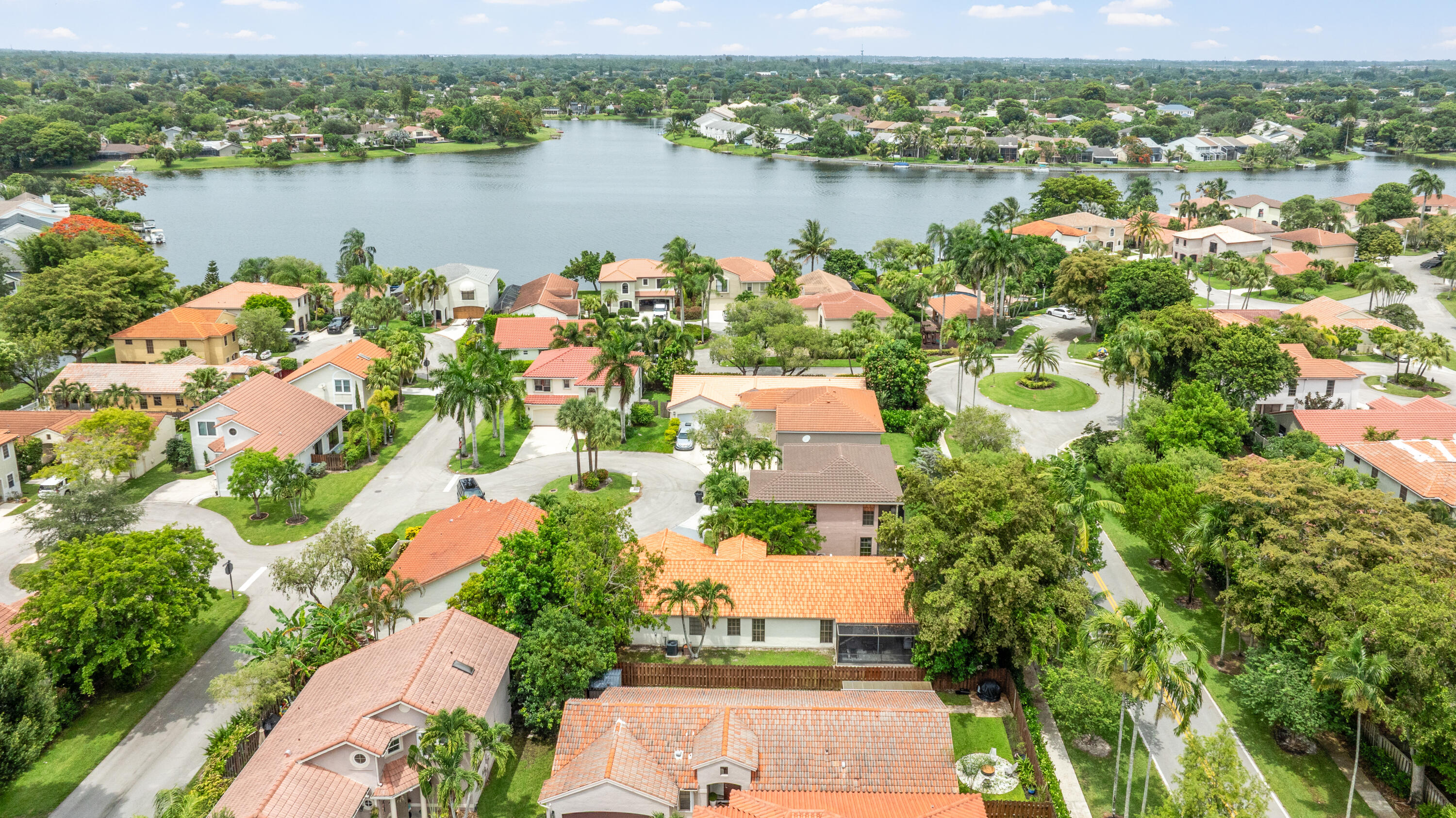 Aerial neighborhood view of tile-roofed homes and palm trees beside a broad lake.
