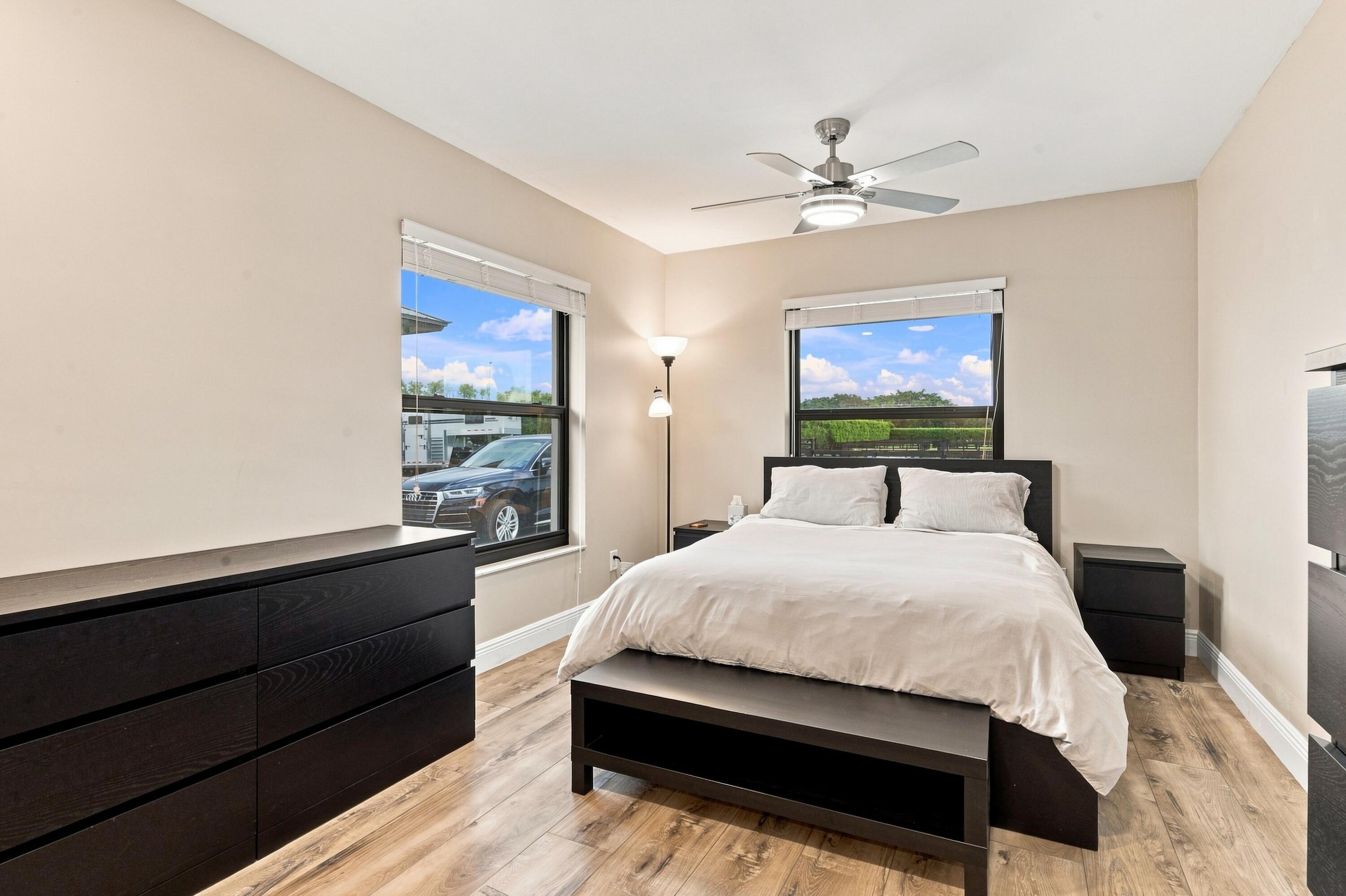 Bedroom with a white bed, dark wood furniture, wood-look floors, and two large windows.