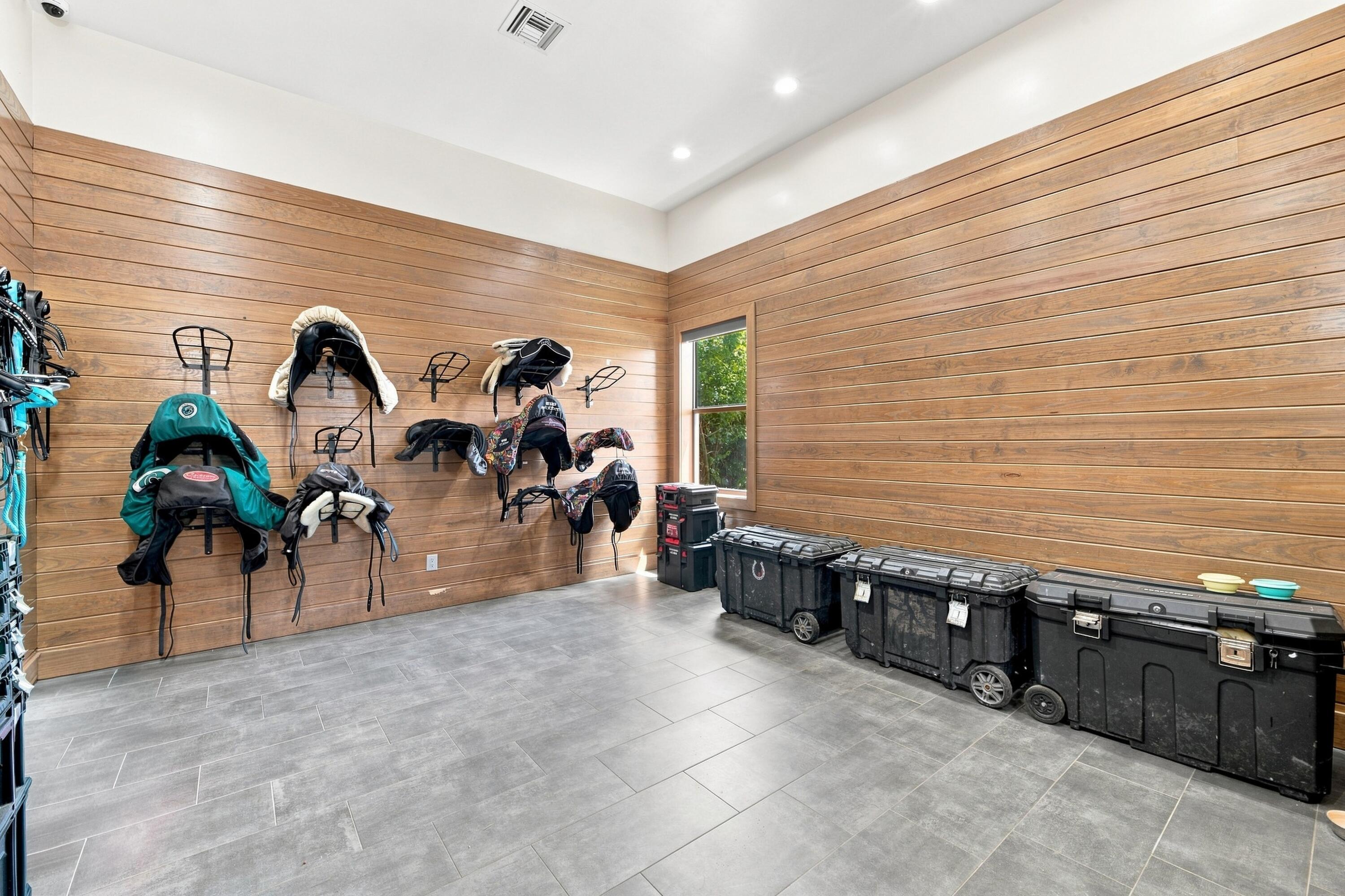 Tack room with saddles mounted on wood-plank walls and storage trunks along the tiled floor.