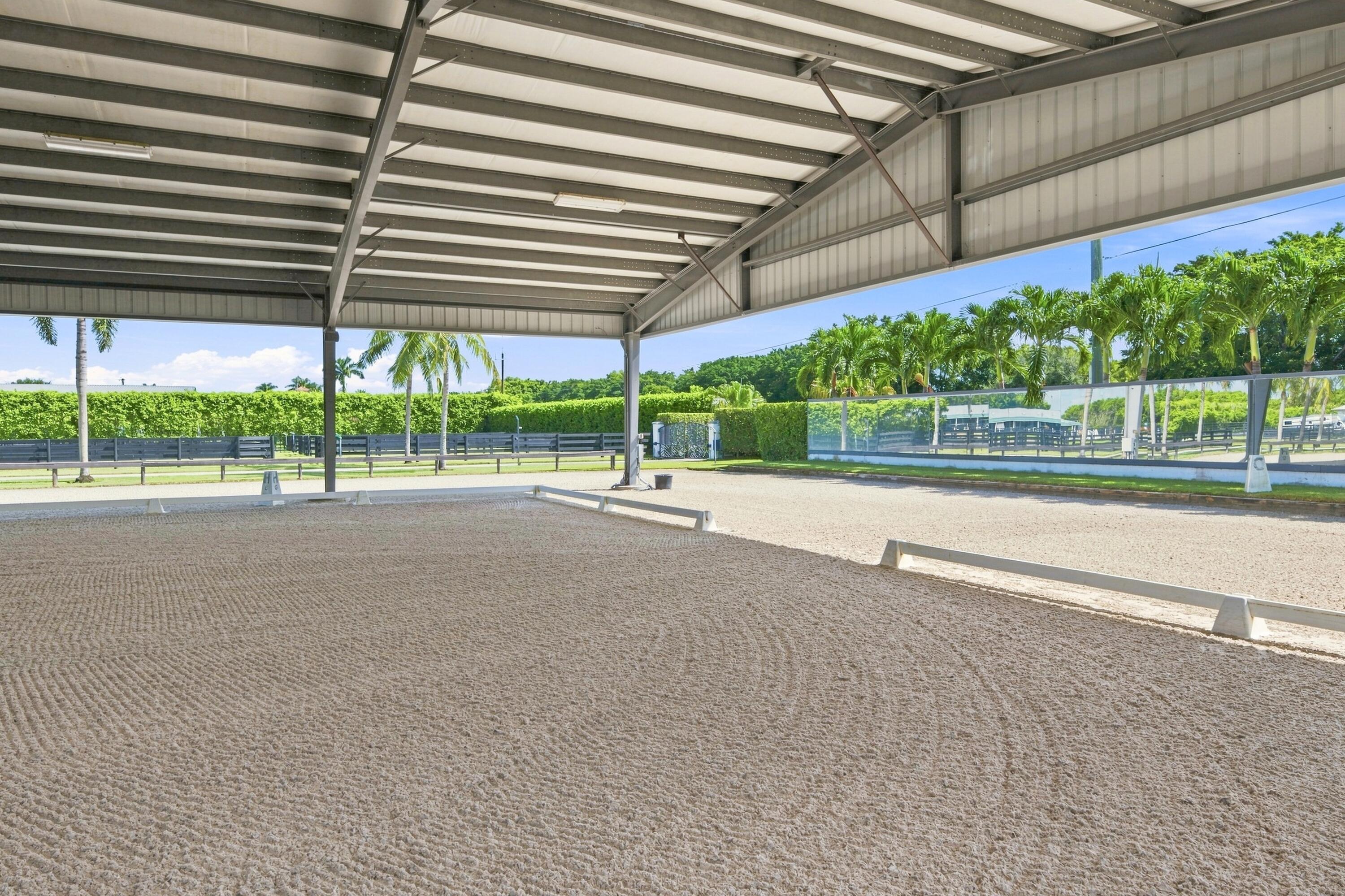 Covered riding arena with groomed sand footing, glass fencing, and palm trees beyond.