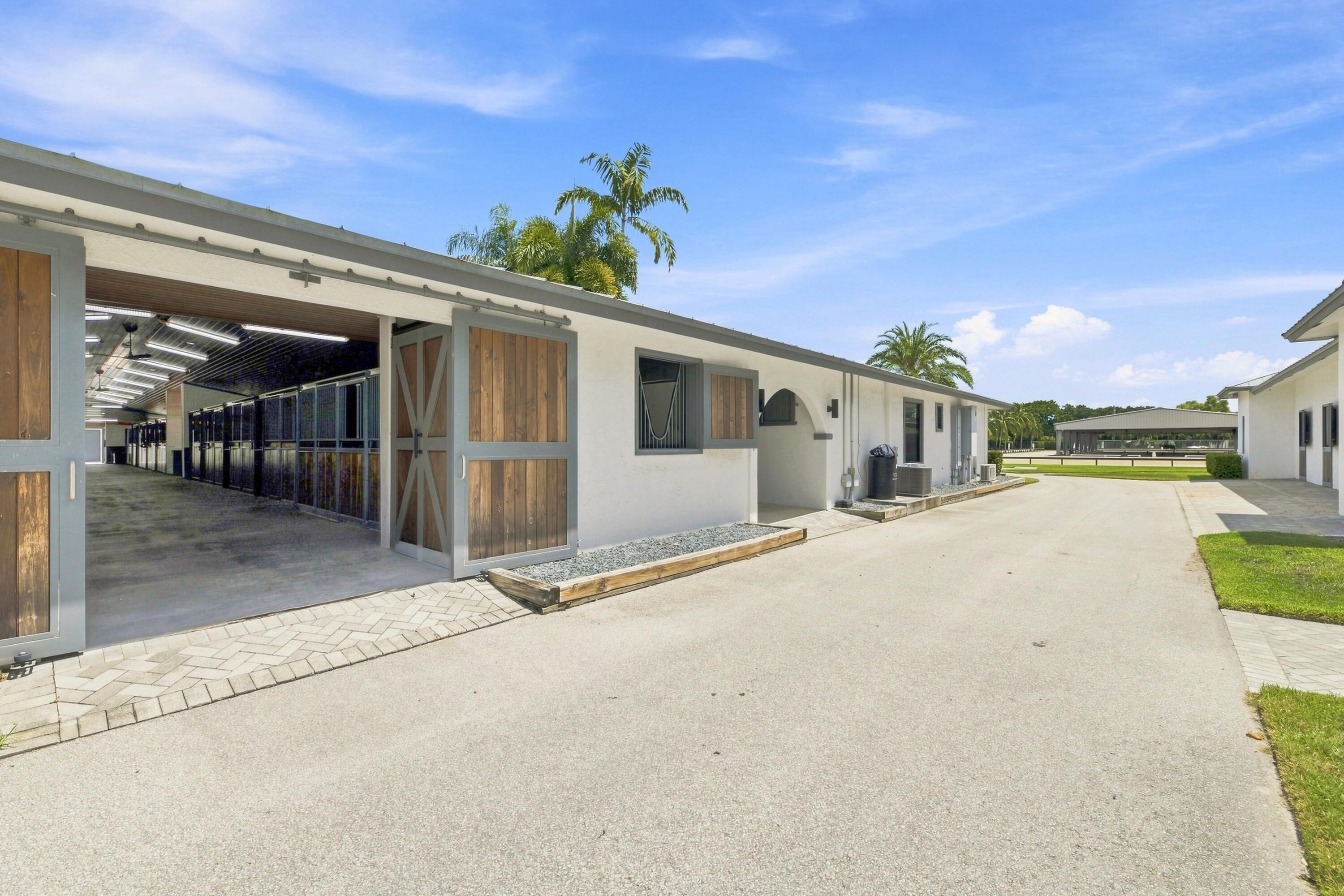 Equestrian stable with open wooden stall doors and a covered riding arena in the distance.