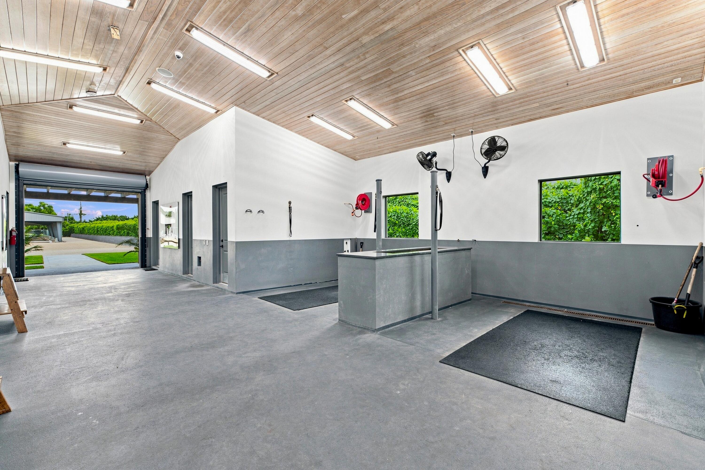 Equestrian wash bay with a central stall, wall-mounted hoses and fans, and an open roll-up door.