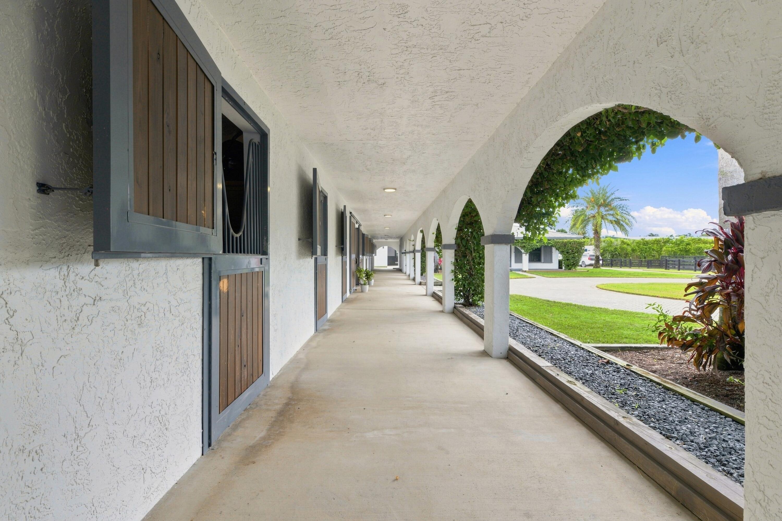 Stable corridor with wooden stall doors and white arches opening onto a sunny lawn.