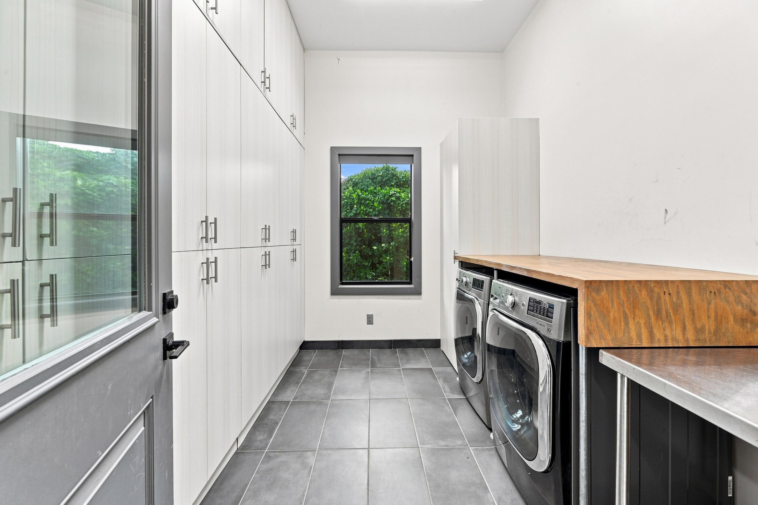 Laundry room with tall white cabinets, gray tile, front-loading machines, and a long wooden countertop.