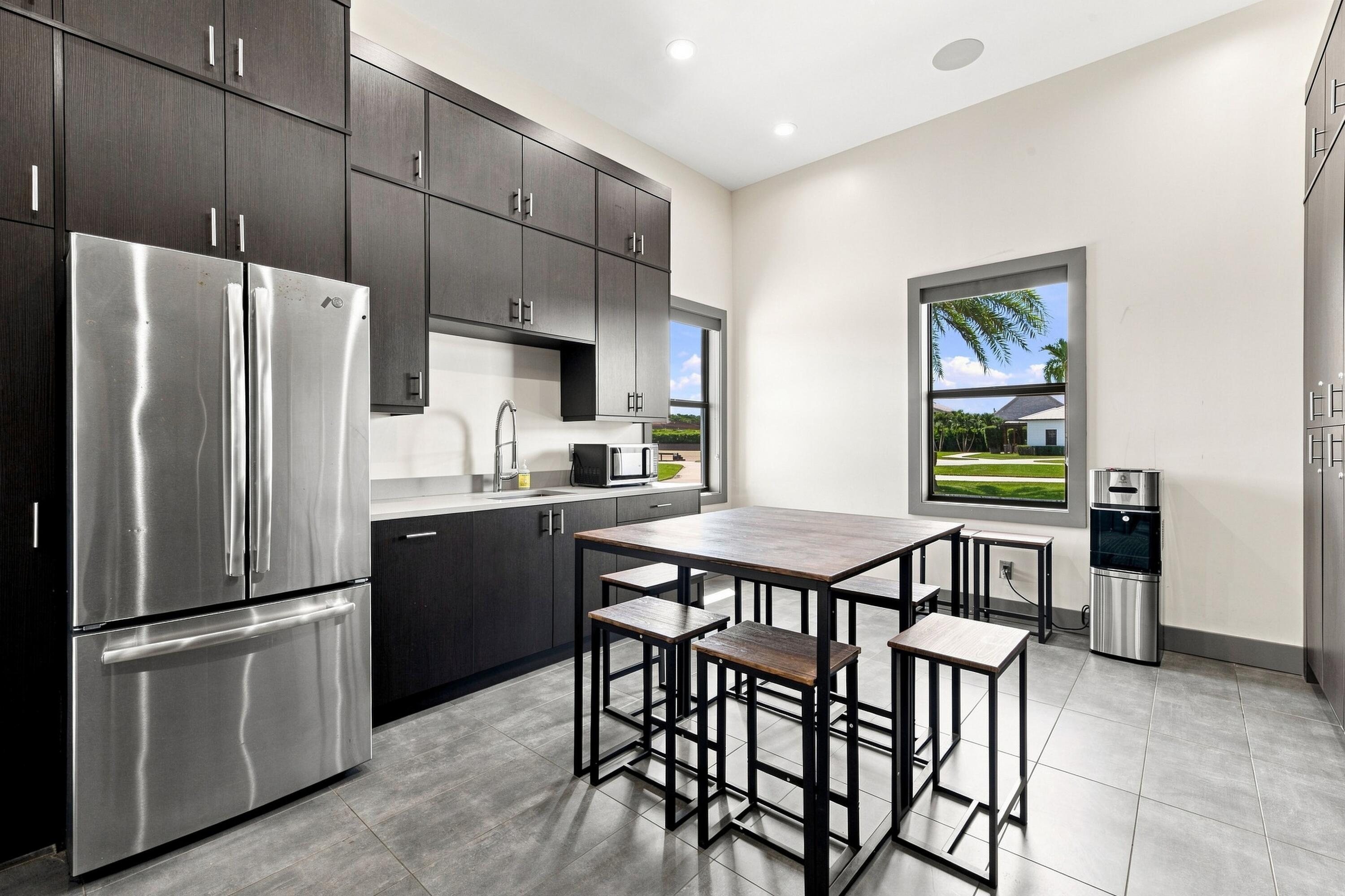 Modern kitchen with dark cabinets, stainless refrigerator, and a table with stools beside two windows.
