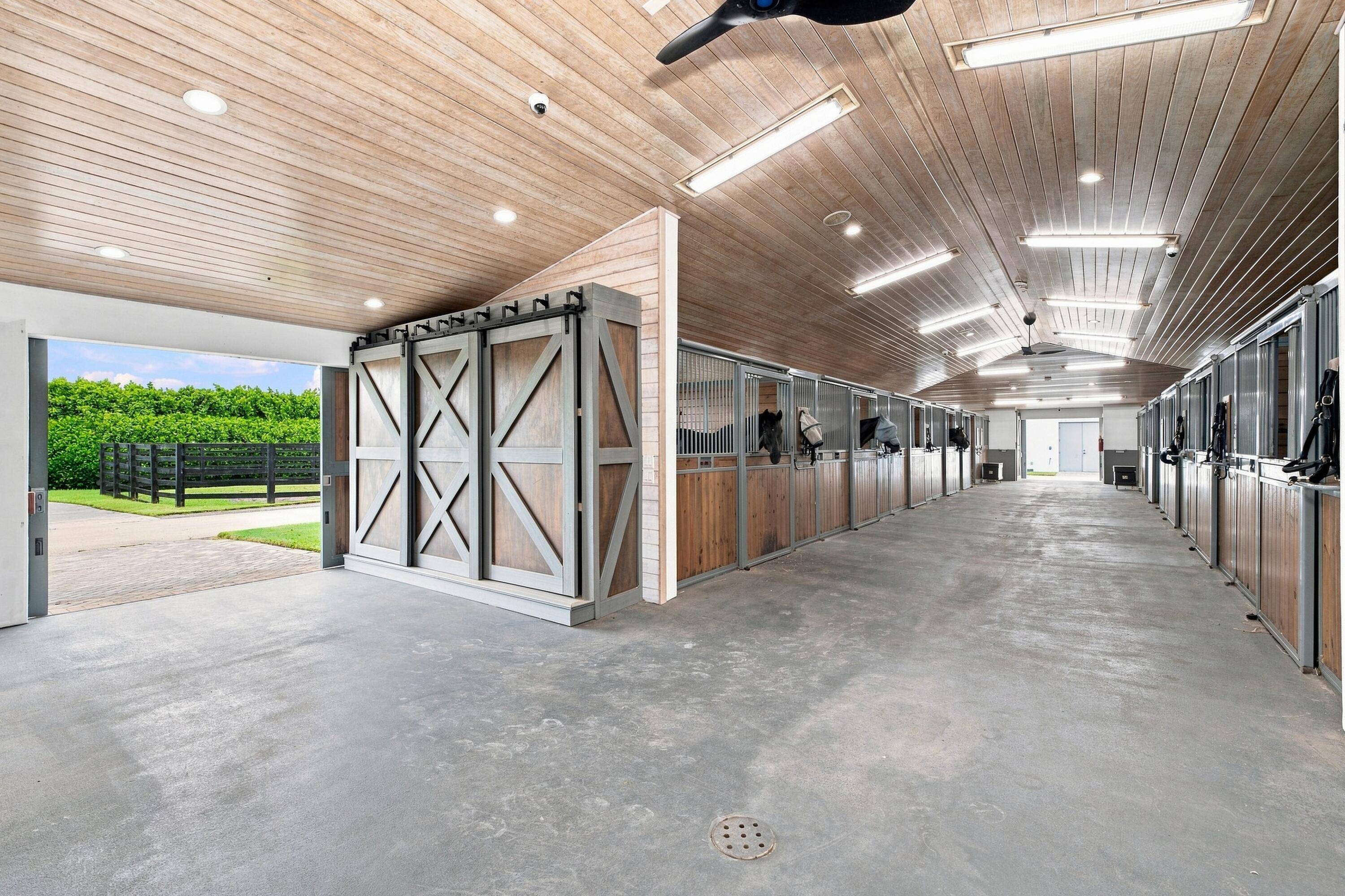 Stable aisle with rows of horse stalls, horses, and a large open barn door.