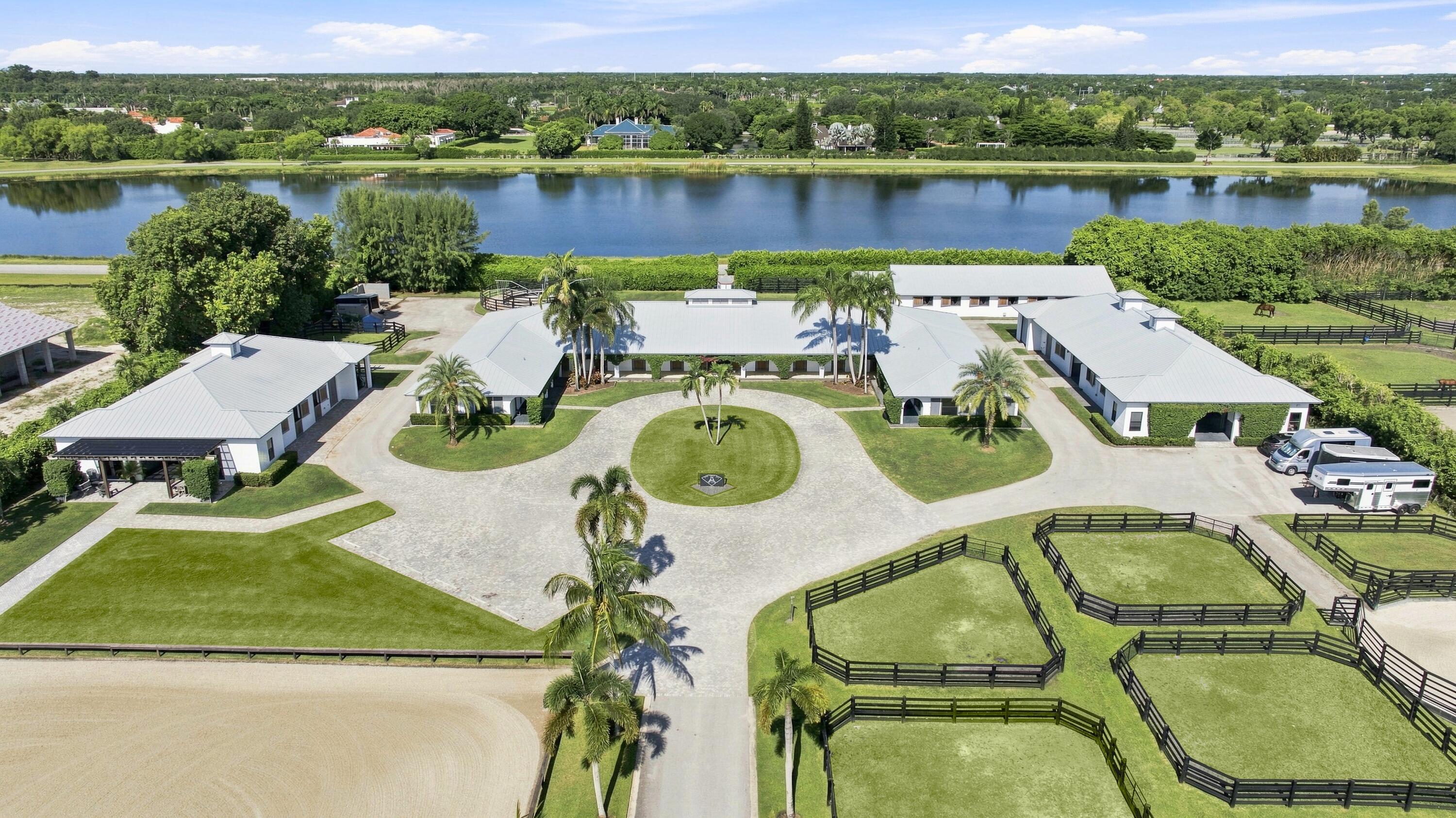 Aerial view of an equestrian estate with white-roofed buildings, fenced paddocks, and a lake.