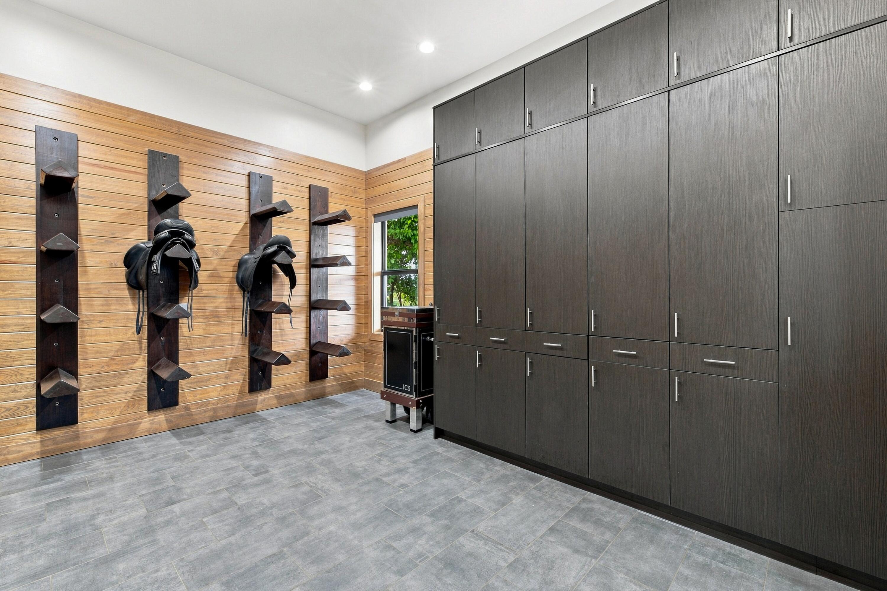 Tack room with dark built-in cabinets and saddles mounted on a wood-plank wall.