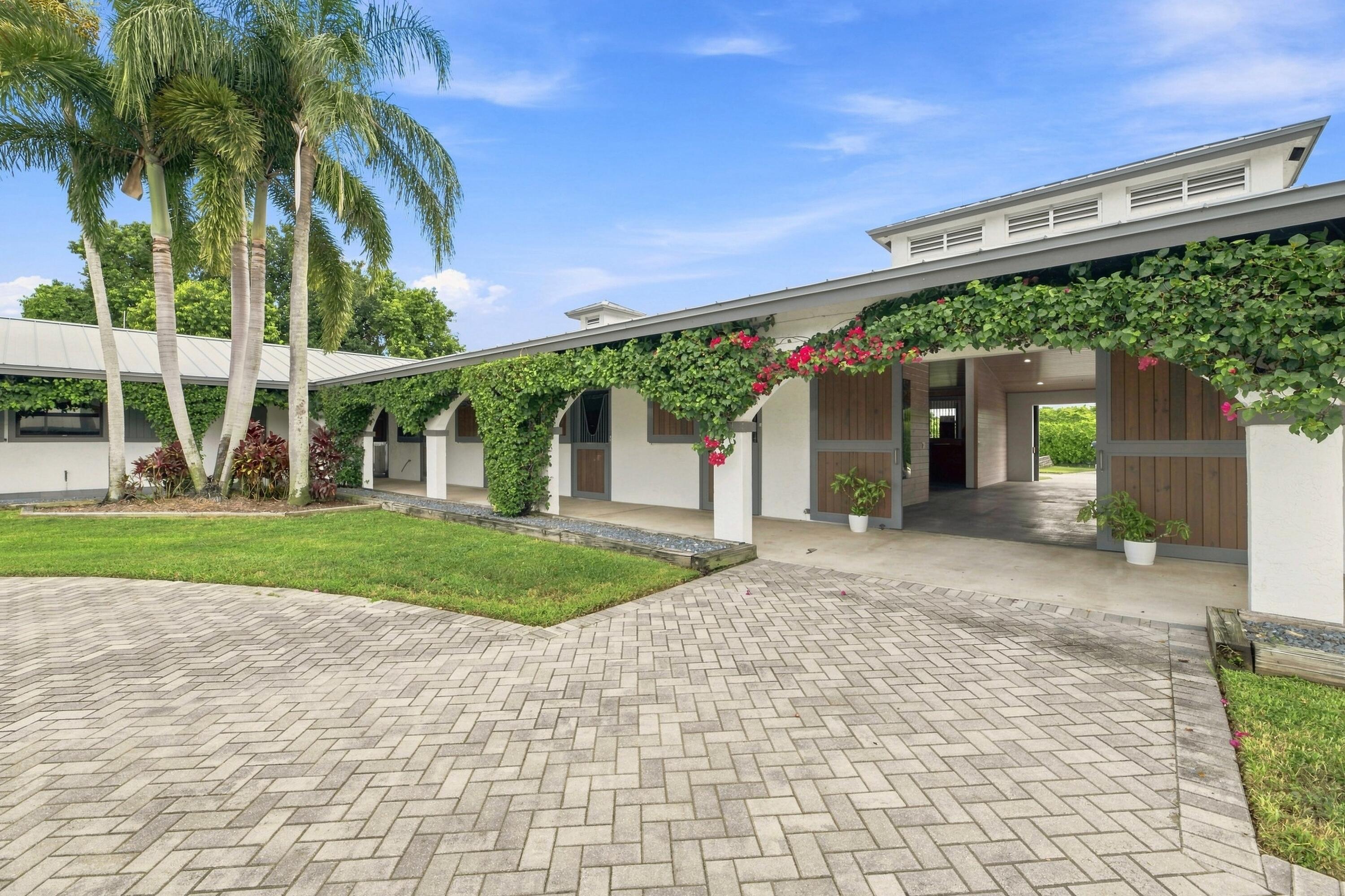 White building with wooden stall doors, flowering vines, palm trees, and a paved courtyard.
