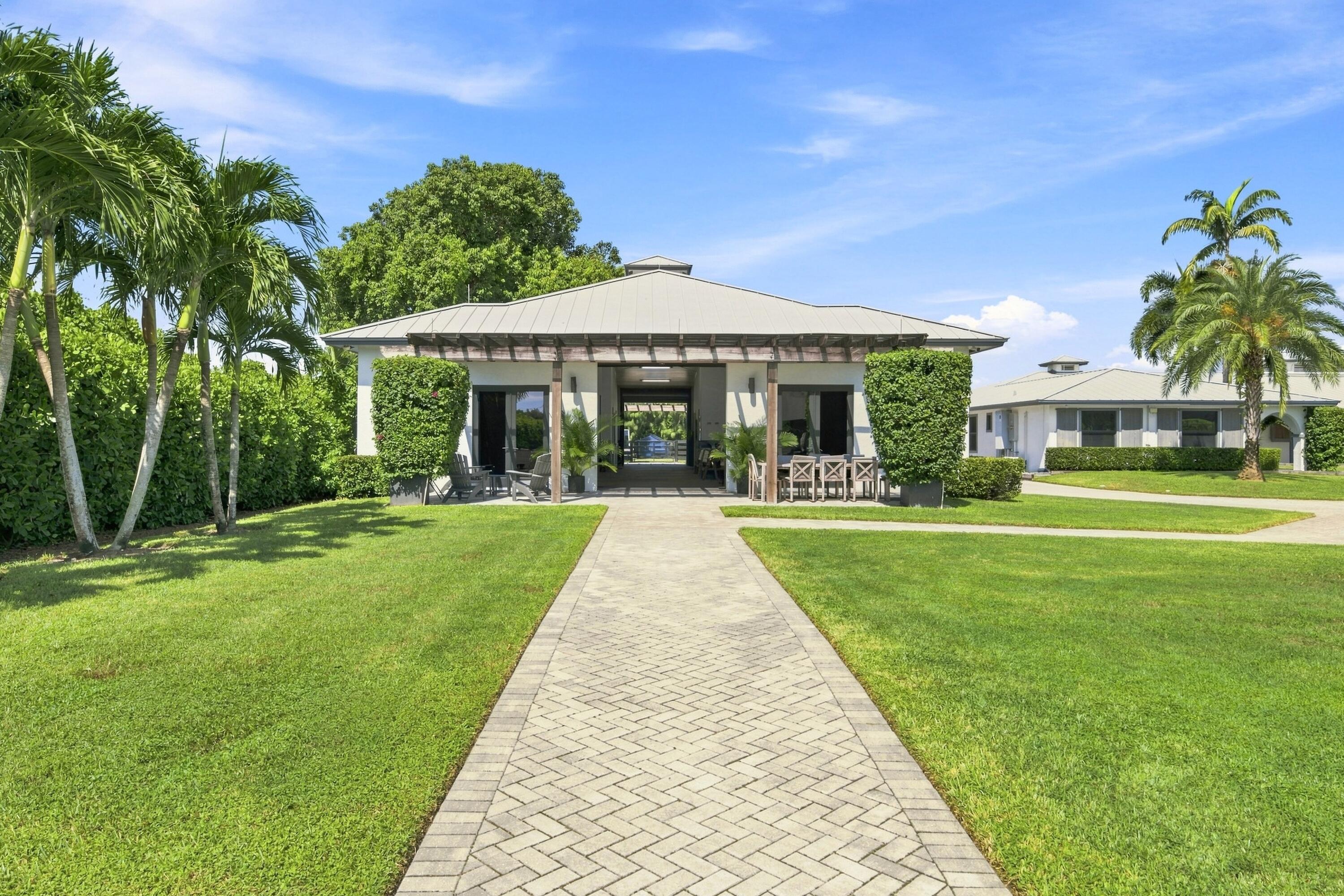 Modern low building with a covered, furnished entrance, paver walkway, manicured lawn, and palm trees.