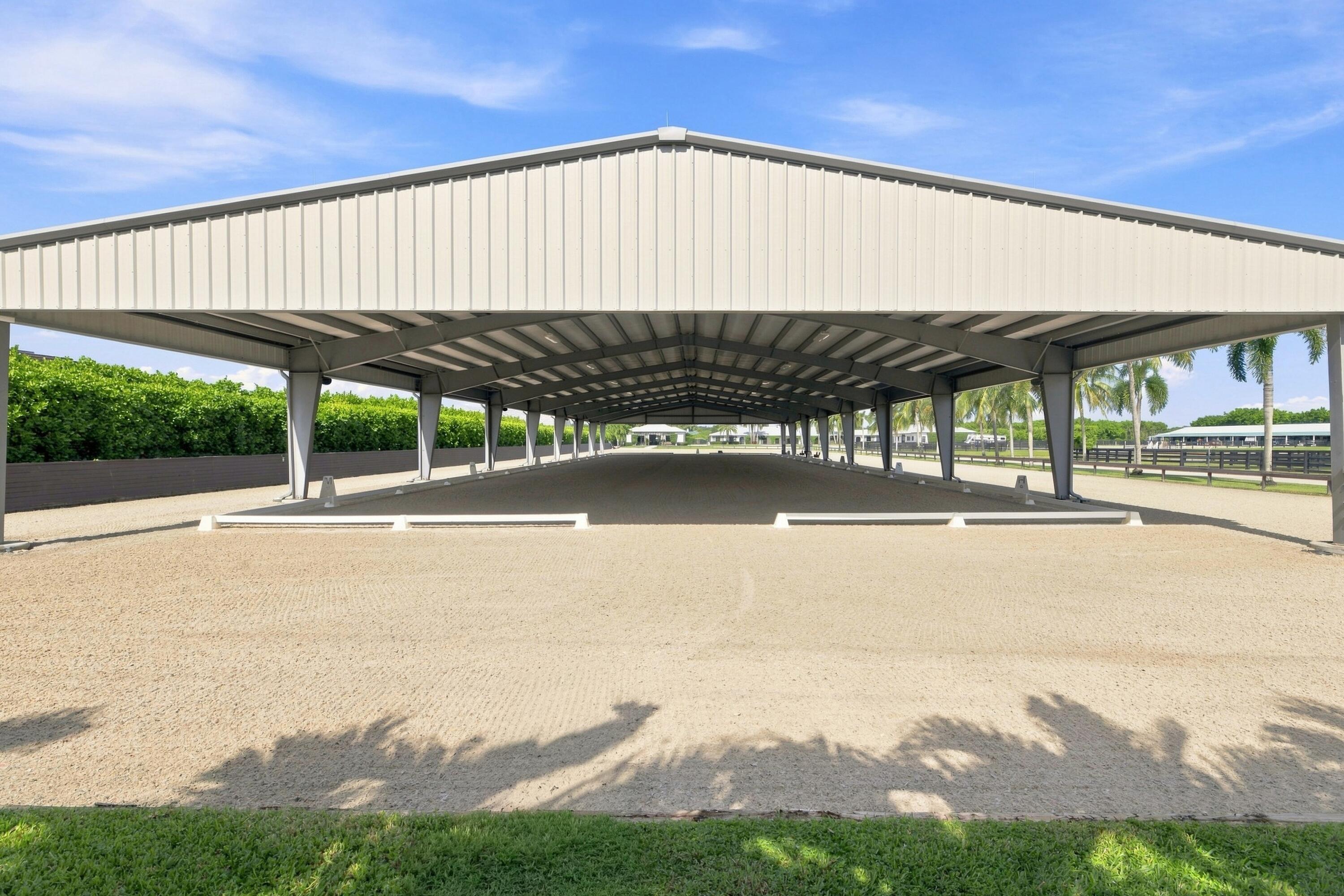 Covered riding arena with a broad sandy surface, white metal roof, and palm trees beyond.