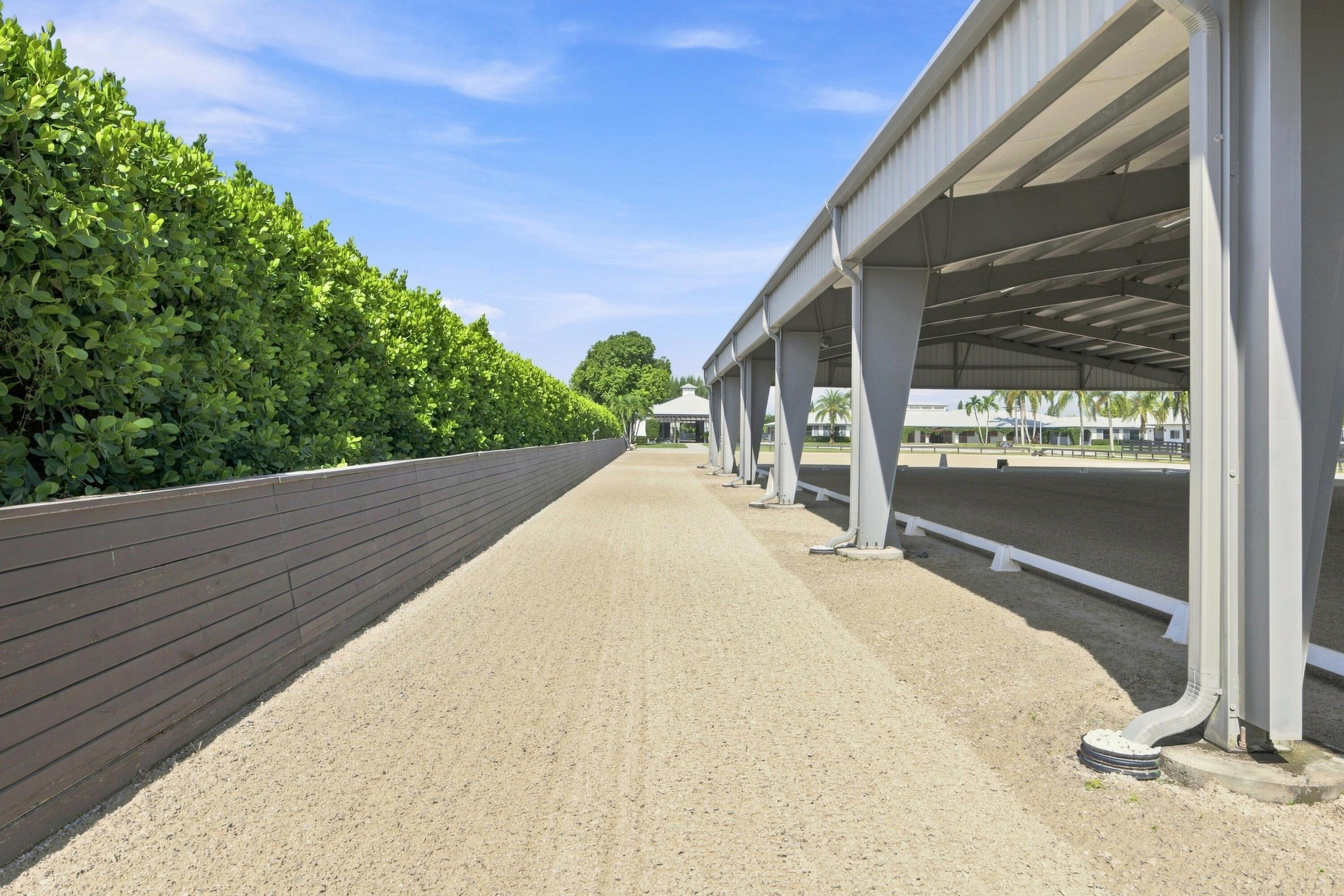 Covered riding arena beside a sandy path, wooden fence, and tall hedge.