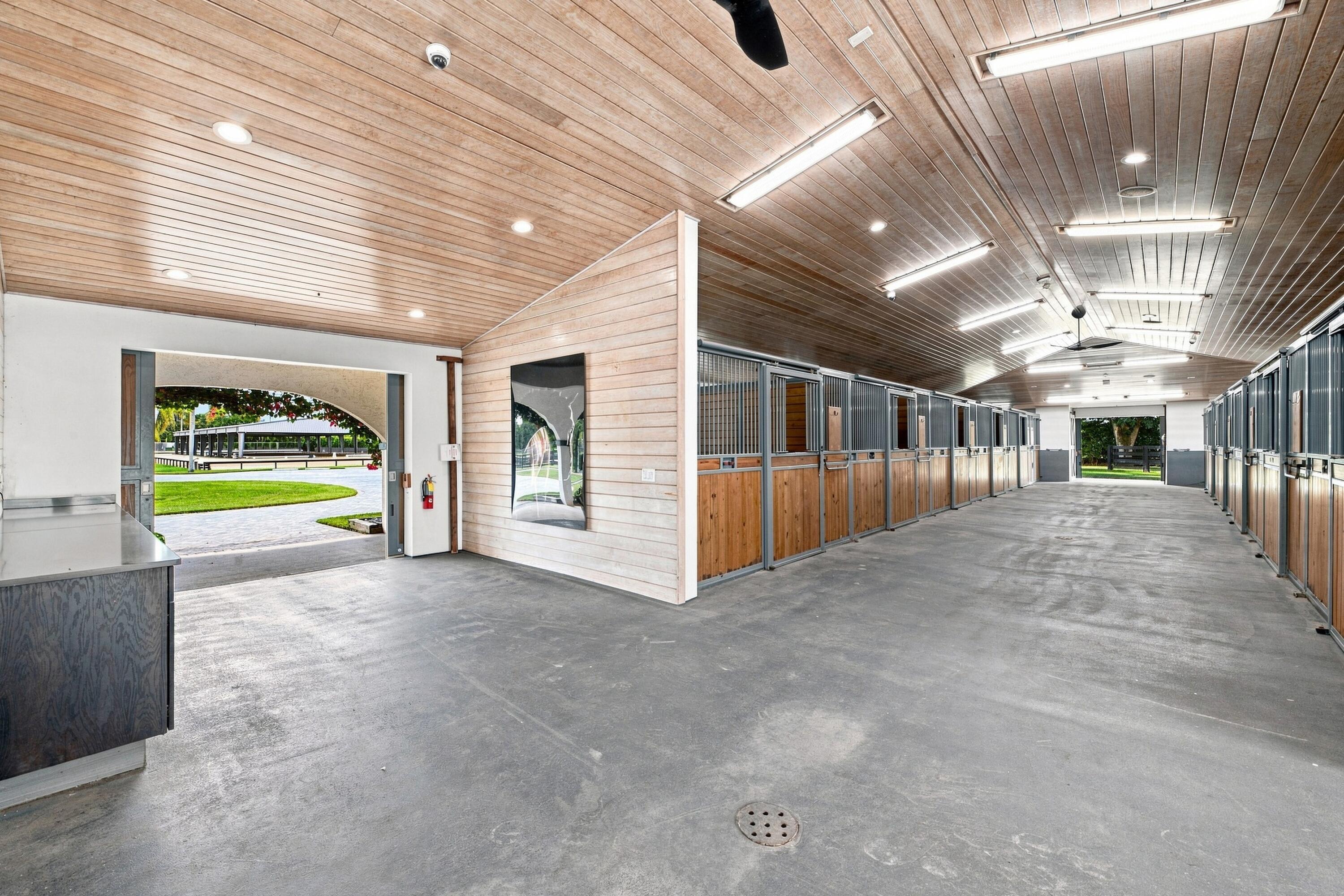 Horse stable interior with rows of wood-and-metal stalls and a wide concrete aisle.