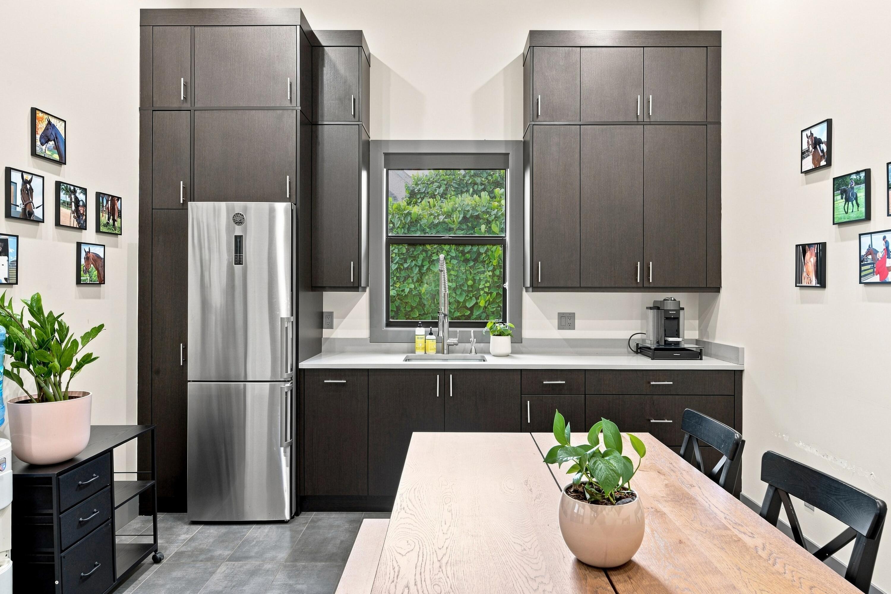 Kitchen with dark cabinetry, stainless steel refrigerator, and dining table before a greenery-facing window.