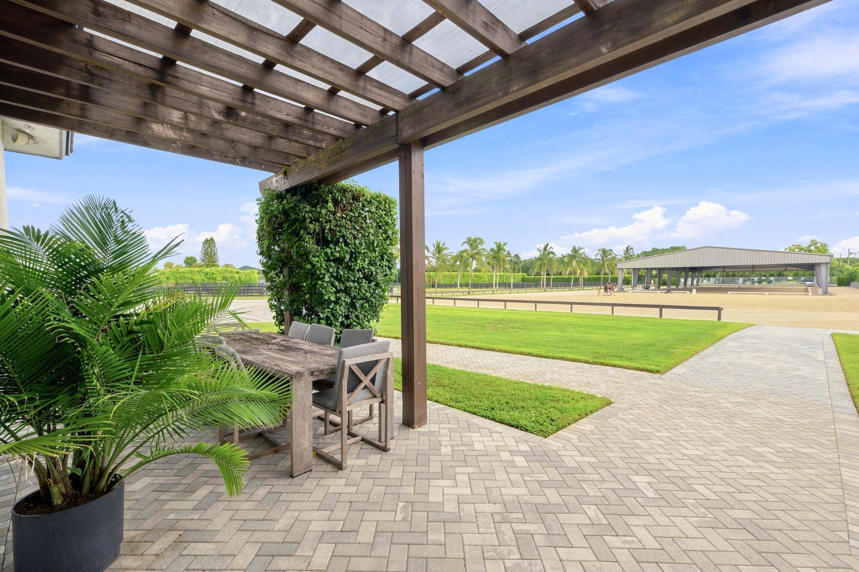 Pergola-covered patio with outdoor dining furniture overlooking a lawn and equestrian arena.