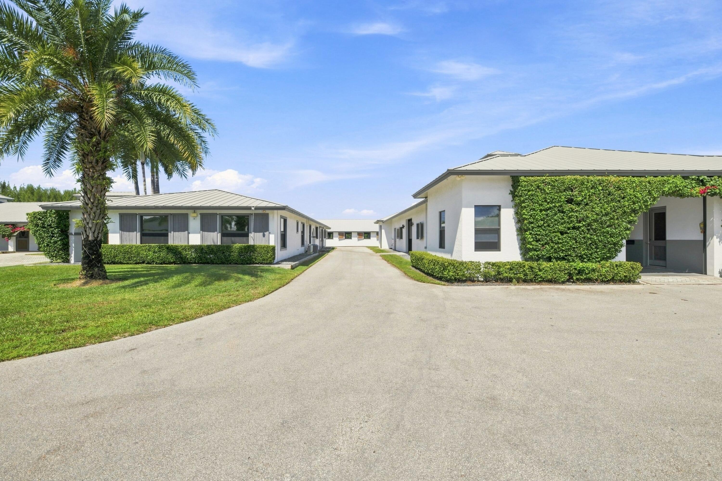 White low-rise buildings line a paved drive beside trimmed hedges and a tall palm tree.