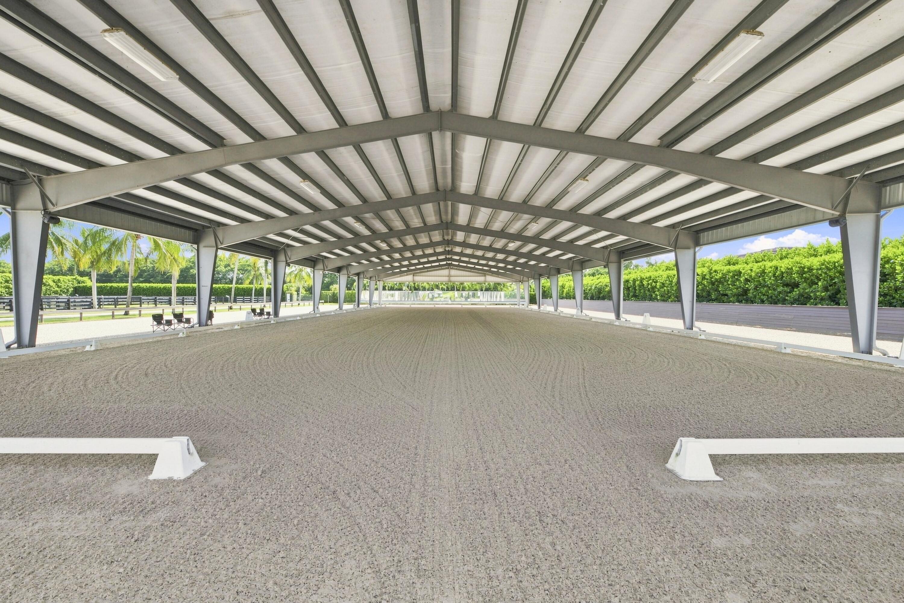 Covered riding arena with groomed sand footing, steel roof beams, and open sides overlooking palm trees.