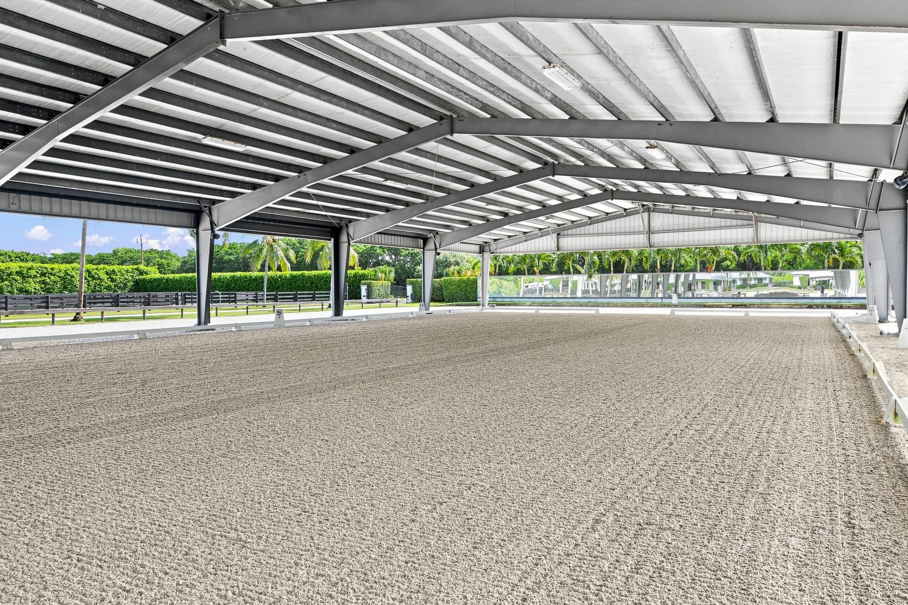 Covered riding arena with groomed sand footing, exposed steel roof trusses, and open sides overlooking palm trees.