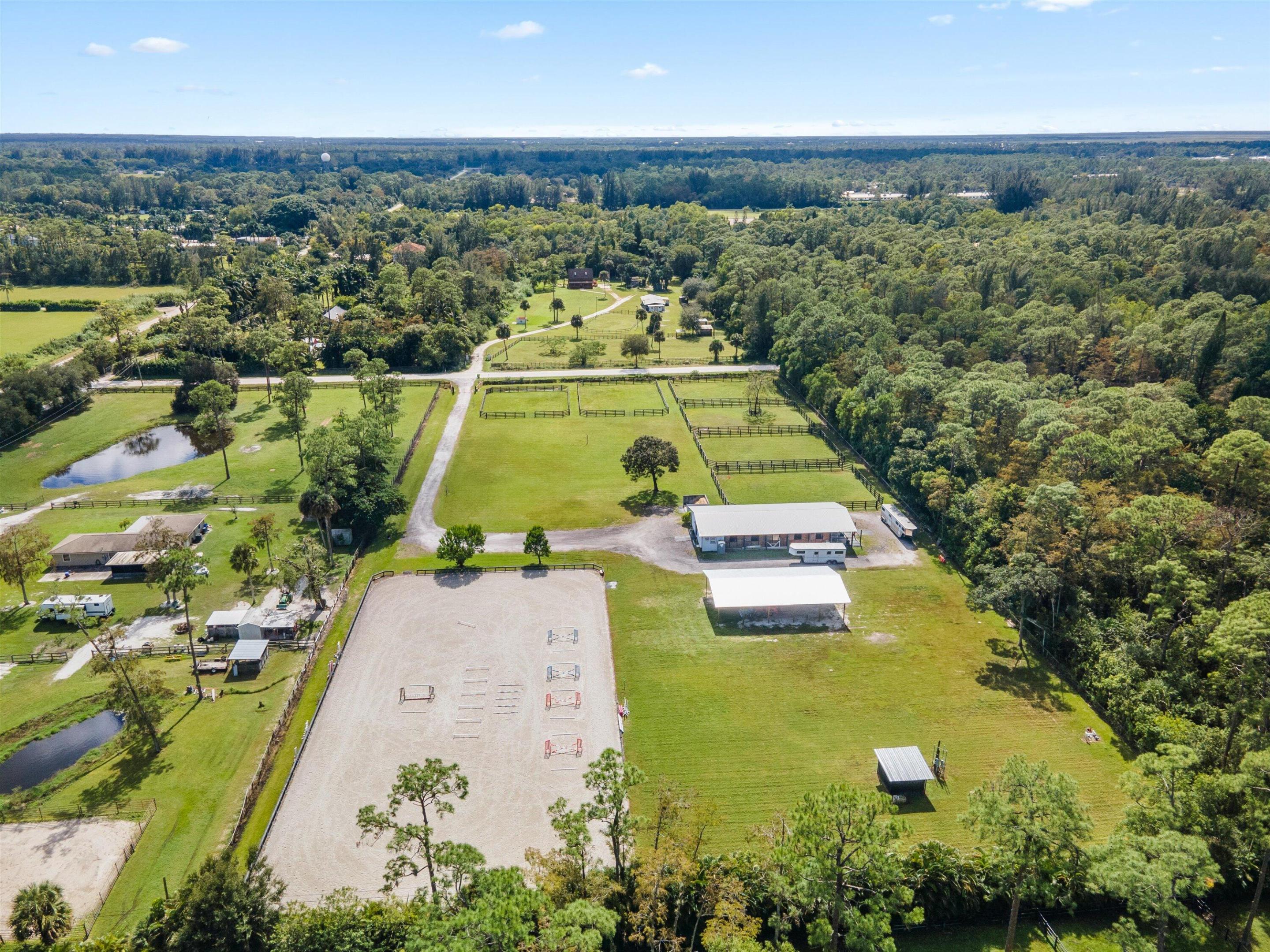 Aerial view of an equestrian property with fenced paddocks, a sand riding arena, and surrounding woodland.