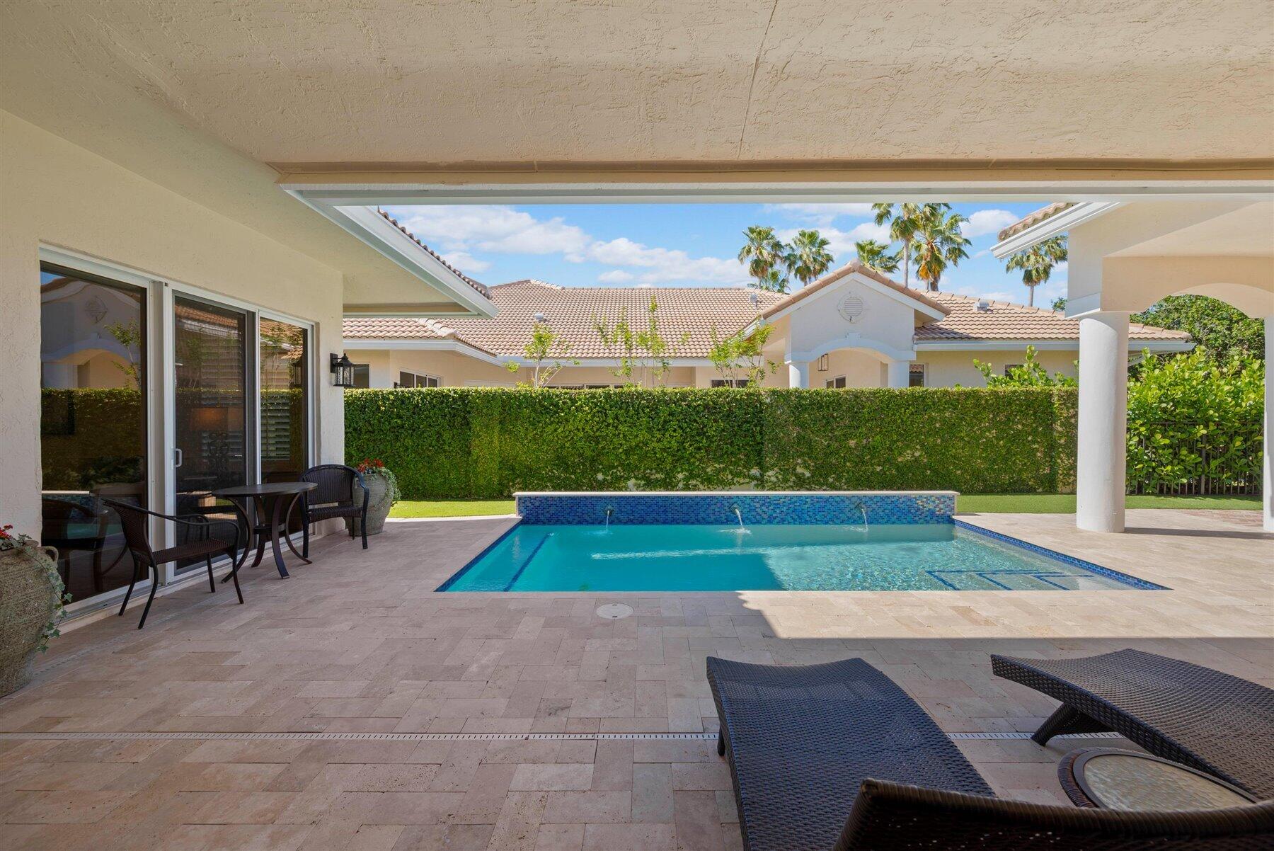 Backyard pool with water jets, covered patio seating, and a tall privacy hedge.