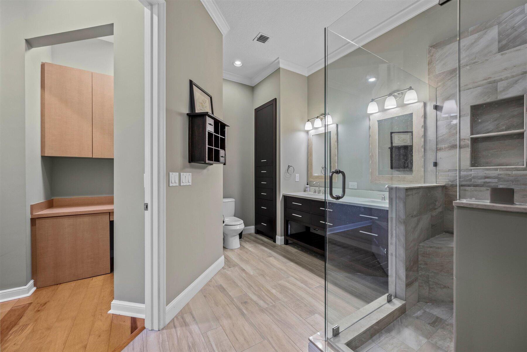 Bathroom with glass-enclosed tiled shower, dark double vanity, and adjacent dressing cabinetry.